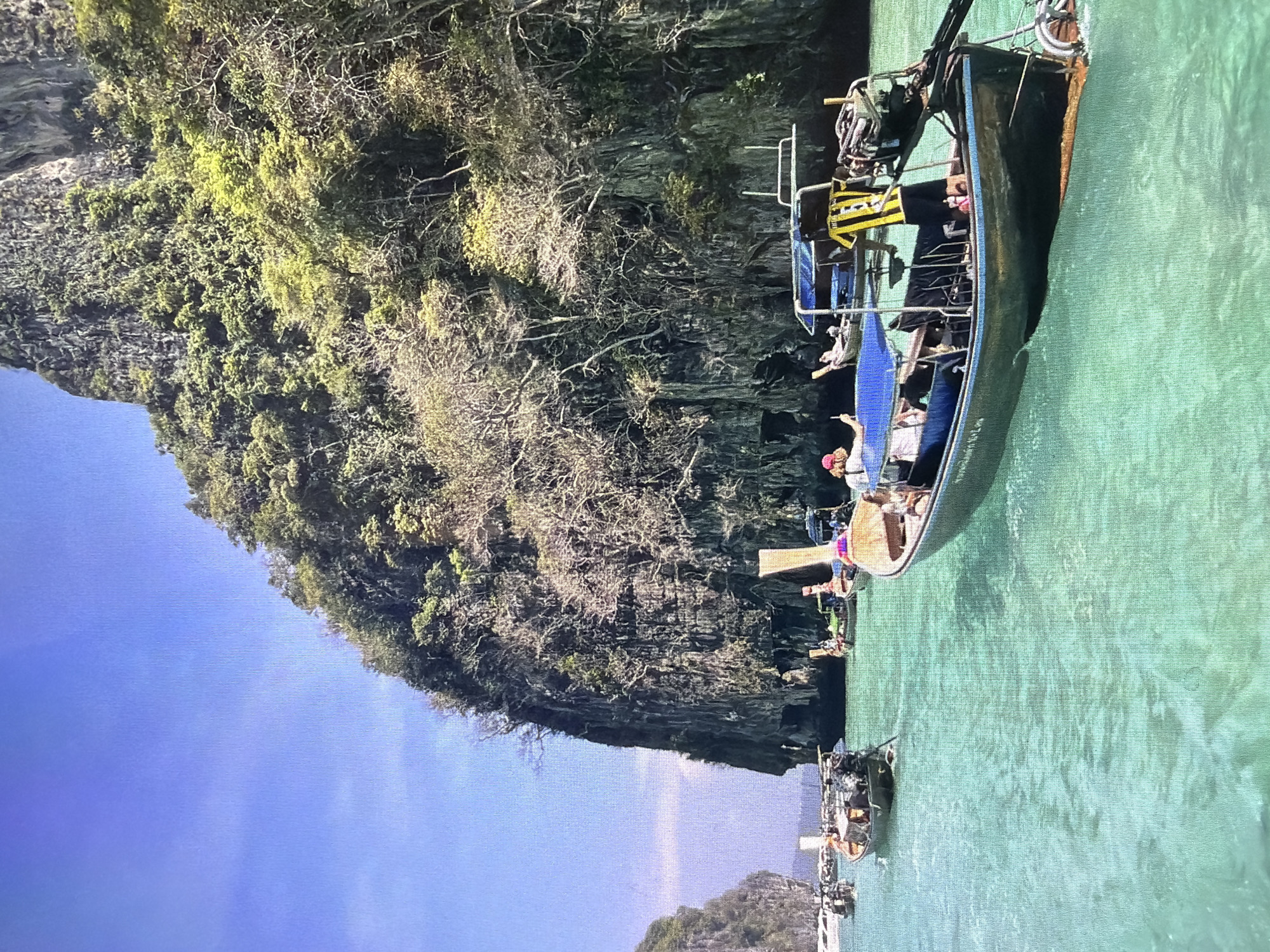 A longtail boat navigates calm, turquoise waters alongside towering limestone karsts covered in dense green and dried vegetation. The image is taken from a slightly elevated perspective, looking towards the front of the boat and the landscape. The sky above is a clear, bright blue.
In the foreground, a dark-hulled longtail boat, typical of Southeast Asian waters, occupies the right side of the frame. Several people are on board; one individual wearing a red hat stands near the front, facing left, seemingly looking out at the scenery. Another person is seated further back, and other passengers are visible in the middle section of the boat. The boat has blue railings and a distinctive yellow and black fabric covering what appears to be part of the engine or canopy structure towards the stern. Ropes and equipment are stored on the boat.
The imposing limestone karsts, characteristic of regions like Thailand or Vietnam, dominate the background. Their steep, rugged surfaces are heavily vegetated, with various shades of green foliage and some areas of bare, dry branches. In the distance, other longtail boats are visible on the water, and more islands or karsts can be seen under the bright blue sky. The water is clear enough to show subtle patterns of the seafloor in some areas.
The scene depicts a sunny day, likely mid-day, with strong natural light. There are no visible signs of specific text or branding. The overall impression is one of a scenic and leisurely boat trip in a tropical environment.