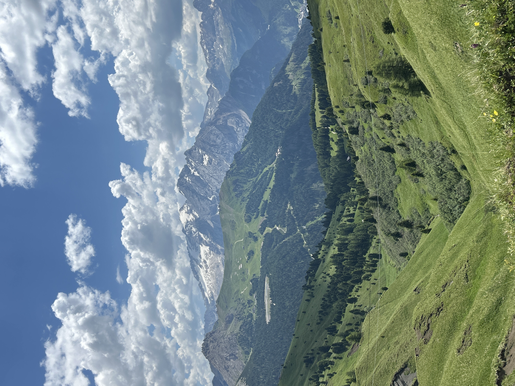 A vertical, medium shot captures a lush green mountainous landscape under a blue sky dotted with white clouds. The foreground consists of rolling green hills with scattered trees and a narrow valley winding through them. In the mid-ground, dense green forests cover the slopes, with a visible lake or reservoir nestled within. Further in the background, majestic mountains rise, their peaks capped with snow, under a dynamic cloudy sky. The scene appears to be a scenic natural vista, likely in a mountainous region during daytime.