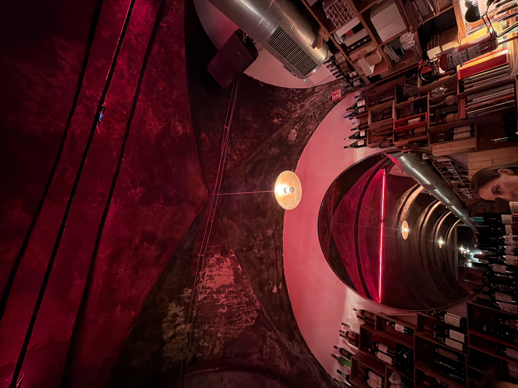 The image shows the interior of a wine bar or cellar in Milan, Italy, with a distinctive arched ceiling illuminated by red lighting. The ceiling has a textured, possibly stone or concrete surface, with visible wiring and some debris. A round, vintage-style pendant light hangs from the ceiling. On the left, there are shelves filled with wine bottles. A large, curved mirror reflects the interior of the establishment, showing more shelves of bottles and the continuation of the arched structure. On the right, more shelves are visible, stocked with bottles and various items, including what appears to be a fire extinguisher. The overall ambiance is dimly lit and intimate, characteristic of a wine cellar or bar. A portion of a woman's face is visible in the lower right corner of the frame.