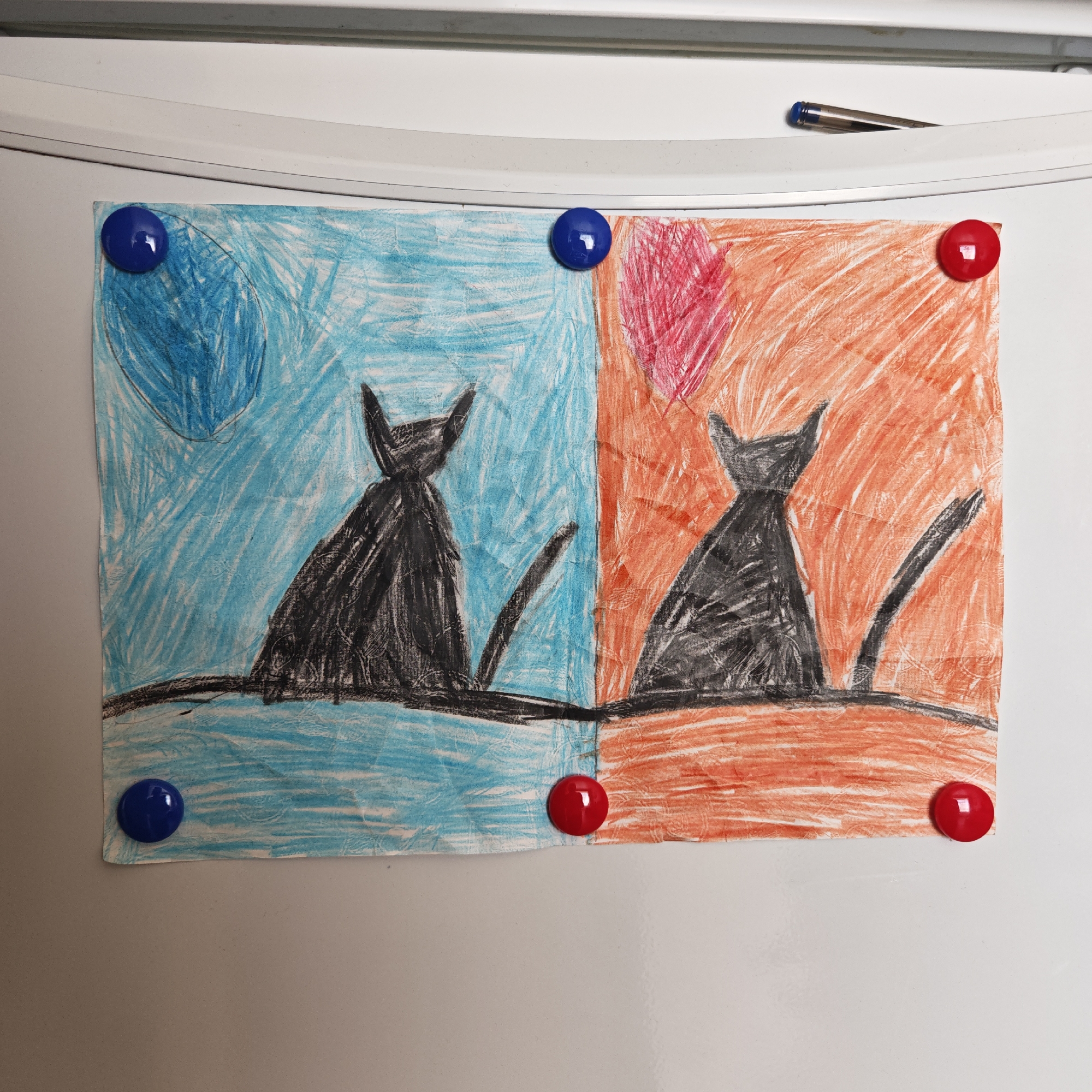 A child's crayon drawing is attached to a white refrigerator with four round magnets. The drawing depicts two black cats, one against a background of blue with a blue circle, and the other against a background of orange with a red circle. The drawing is affixed to the refrigerator with blue magnets in the upper left and lower left corners, and red magnets in the upper right and lower right corners. A pen rests on the top edge of the refrigerator. This scene is likely in a domestic setting, possibly a kitchen in Taganrog City District, Russia, although the location is only inferred from potential source information and cannot be definitively confirmed from the image itself.