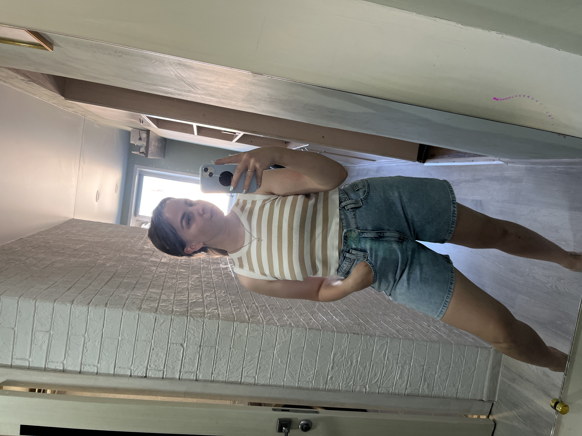 A woman is taking a mirror selfie indoors. She is wearing a striped tank top and denim shorts. The background shows elements of a room, including a white brick wall and a doorframe. There is no information present to confirm the location is Izhevsk, Russia.