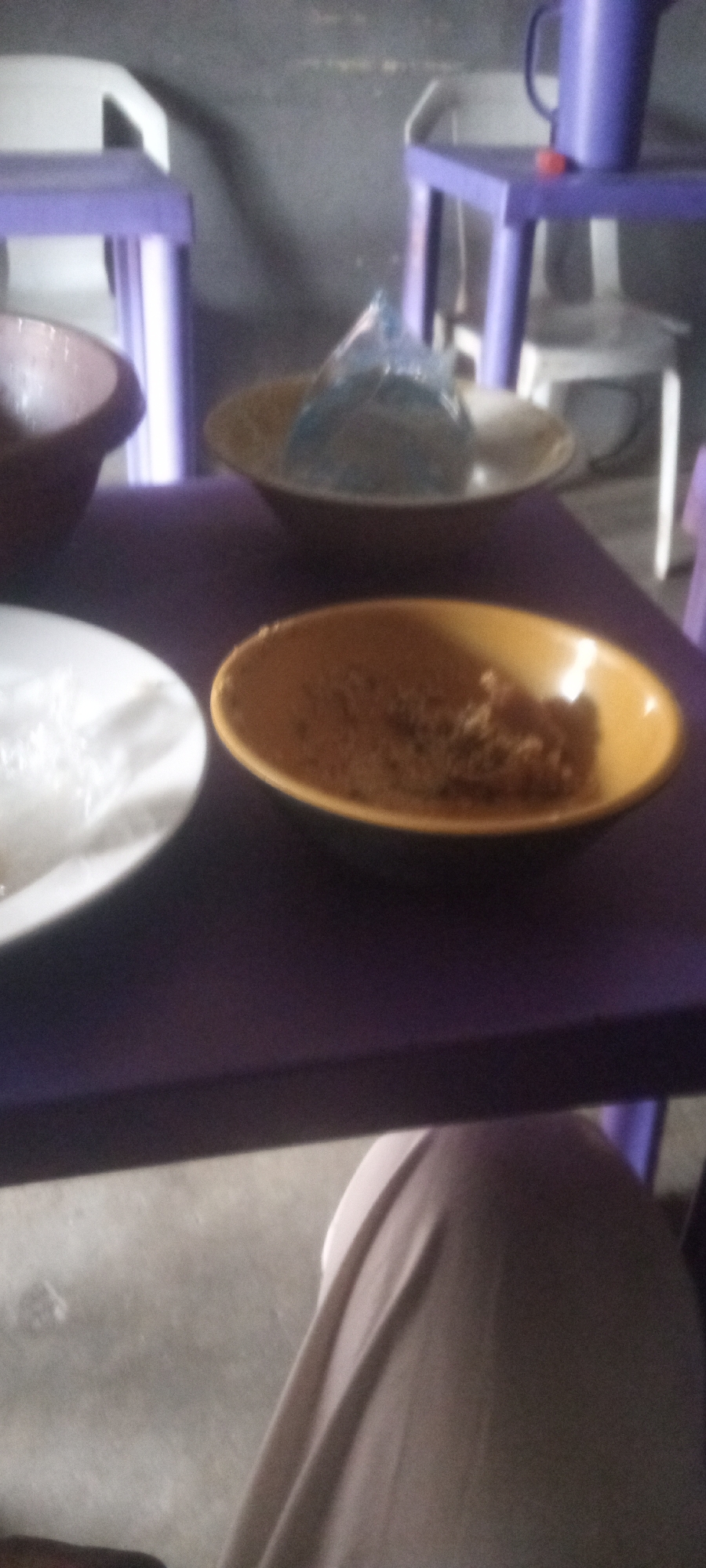 The image depicts a table setting with several dishes. A white plate sits in the lower left corner, alongside a yellow dish containing some food item, and a bowl with something inside, partly obscured. In the background, another dish holds a partially visible plastic bag. The table is purple, and several purple and white chairs are visible. A purple pitcher is also present on a table behind the first. It's unclear where in the city of Unknown, Unknown this is taking place. A person’s tan clothing is visible in the bottom portion of the image.