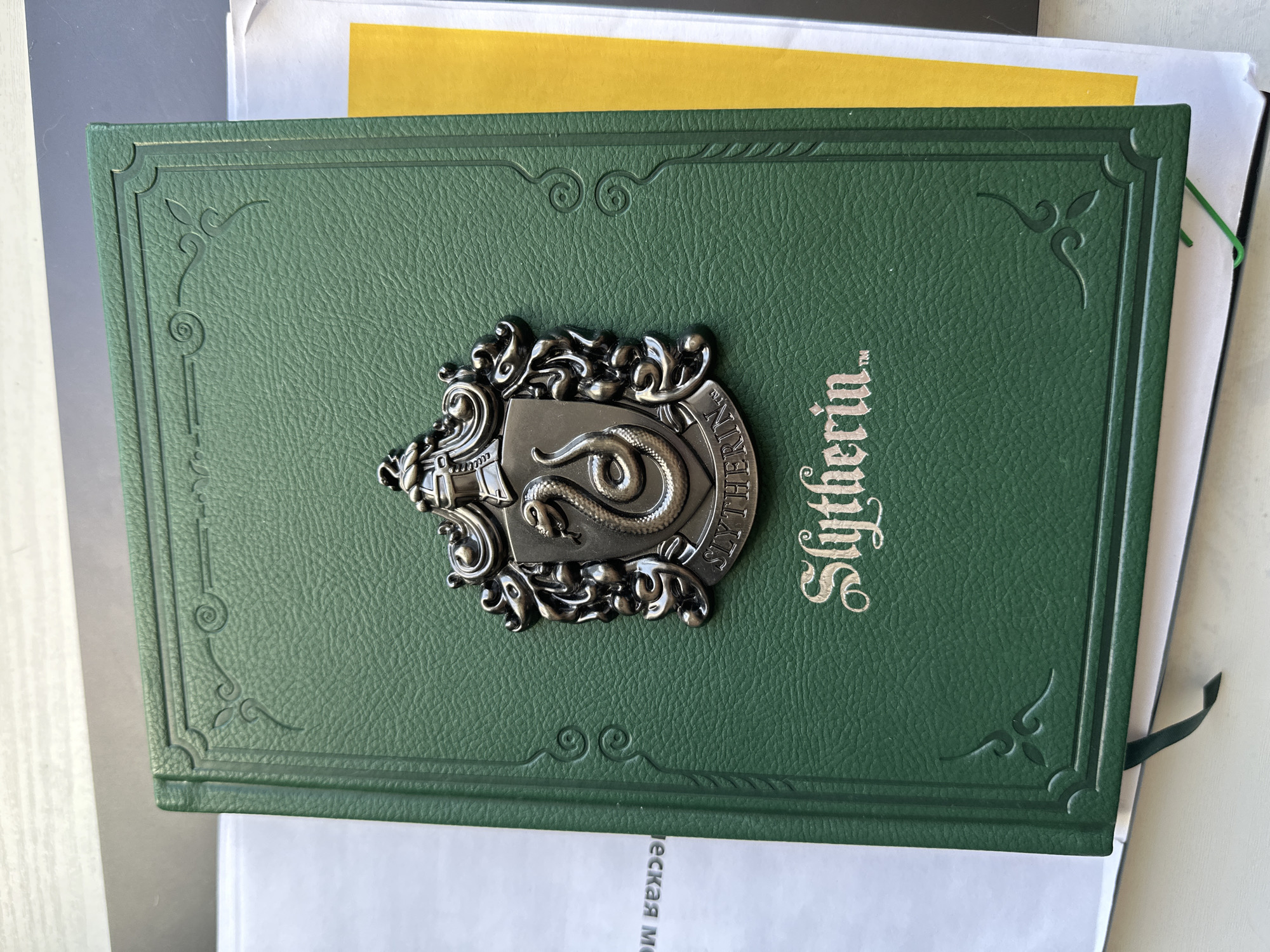The image shows a green book with a silver Slytherin emblem on its cover. The emblem features a snake coiled around a shield. The word "Slytherin" is printed vertically in silver on the book's cover to the right of the emblem. The book is placed on top of several papers, with a yellow sheet visible between some of the pages. The bottom of one of the papers has Cyrillic text on it. No people are visible. The location is not evident from the image.