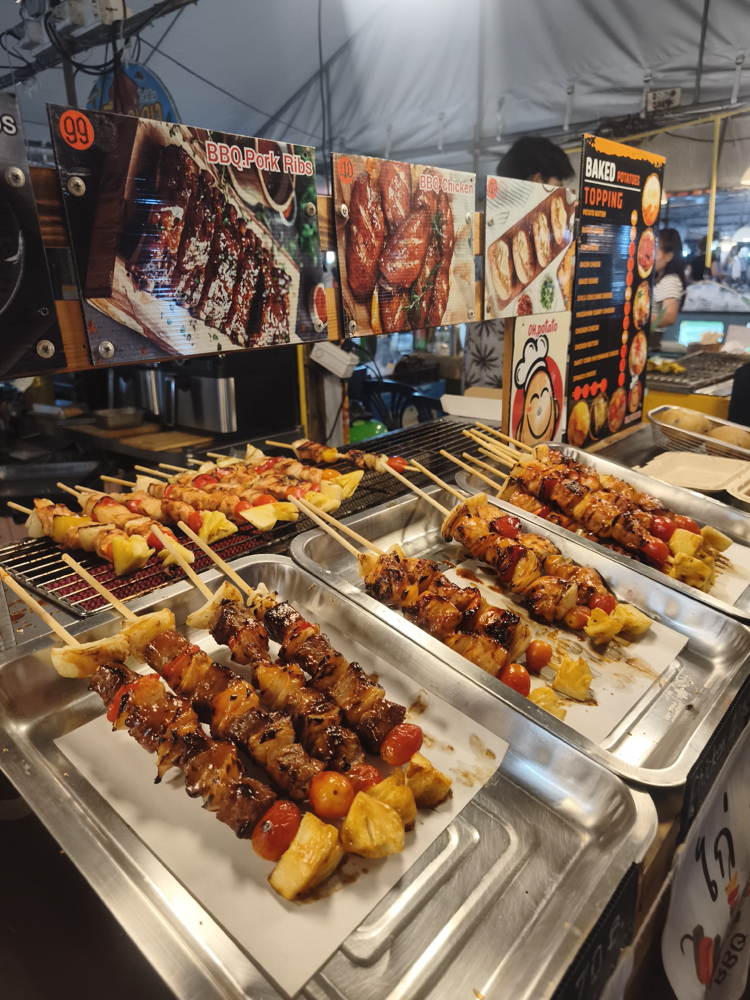 The image shows a food stall selling BBQ skewers and other items in บ้านบางรักษ์, Thailand. In the background are posters advertising "BBQ Pork Ribs" (99), "BBQ Chicken" (40), and "Baked Potatoes Topping." Three metal trays lined with parchment paper are in the foreground. The first tray contains skewers of glazed meat, red bell peppers, pineapple, and tomatoes. The second tray contains similar skewers with what appears to be chicken as the main ingredient. The third tray contains similar skewers, some still on the grill, consisting of chicken or another light meat, pineapple, and tomatoes. A sign with a cartoon potato chef and "oh, potato" is visible. A red sticker indicates the items are BBQ Chicken. A person stands in the background near the "Baked Potatoes Topping" sign.