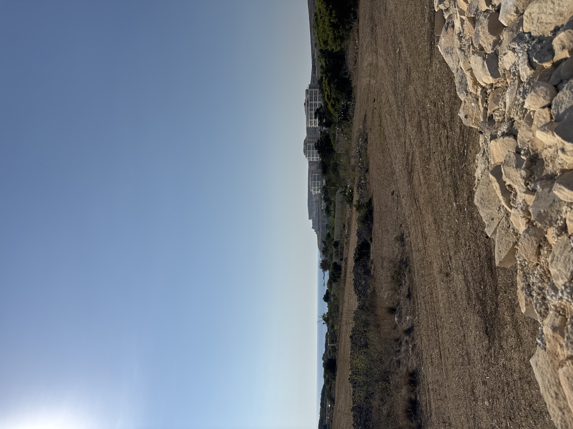 The image depicts a bright, clear day with a vast expanse of blue sky occupying the upper portion. In the mid-ground, a row of modern, white multi-story buildings with rounded edges is visible, set against a backdrop of lush green trees and a distant rocky landscape. The foreground features a dry, cultivated field with rough, brown soil and scattered rocks, leading towards a rocky wall constructed from light-colored stones. The ocean is visible on the horizon, with a calm, blue surface meeting the sky. The sun, a bright white circle, is positioned in the upper left corner, casting a strong light across the scene.