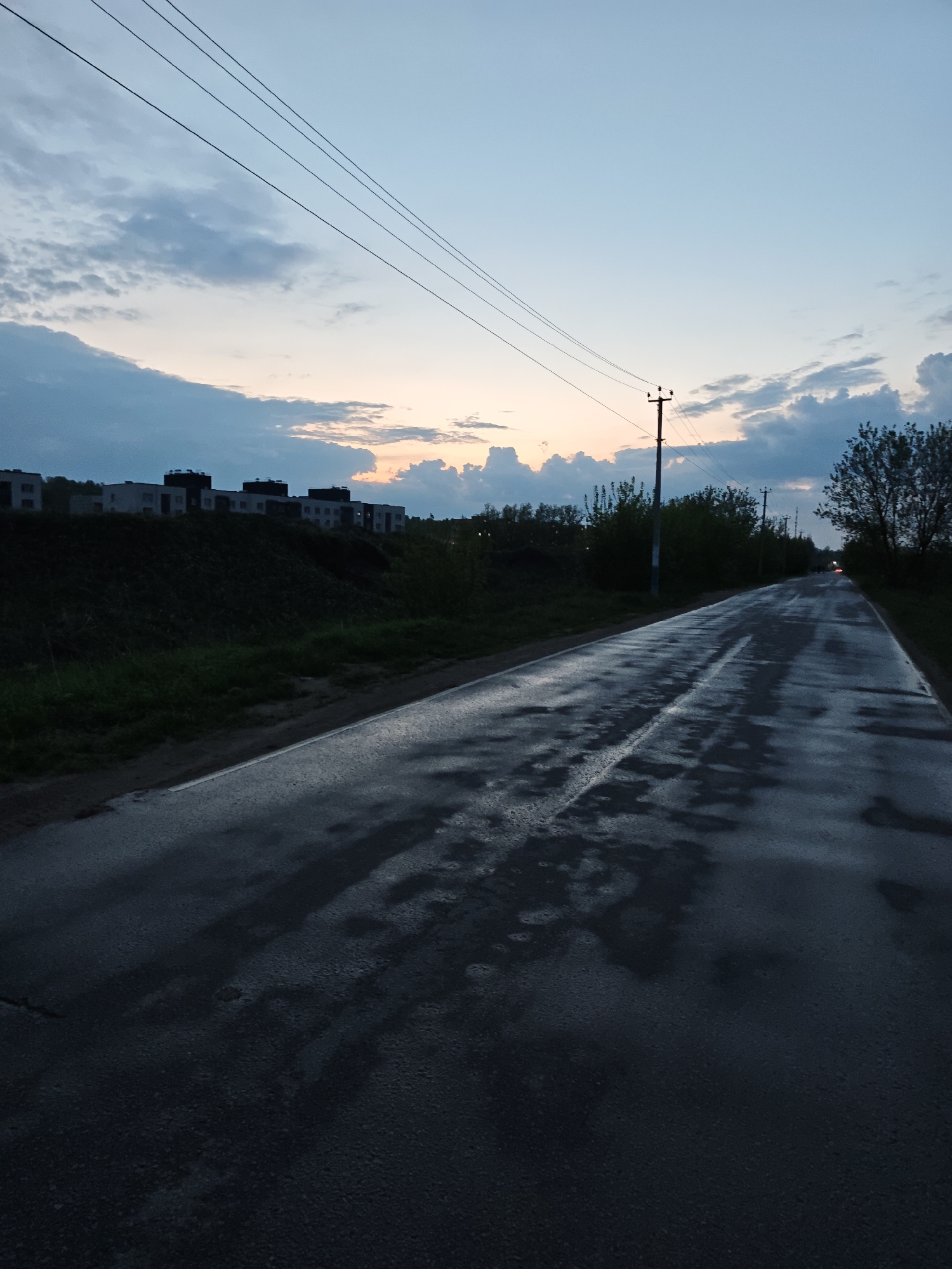 The image captures a wet, dark asphalt road stretching into the distance under a sky transitioning between day and night. The road has a single broken white line marking its center. Patches of standing water reflect the sky and surroundings. On the left, a grassy embankment slopes upwards, topped with a cluster of buildings, likely apartment blocks, silhouetted against the twilight sky. Further along the road, power lines are visible against the sky, connected to a utility pole on the right side of the road. Trees and bushes line the roadside. In the far distance, there is a faint red glow, possibly the taillights of a vehicle. The scene is in Yakhroma, Russia.