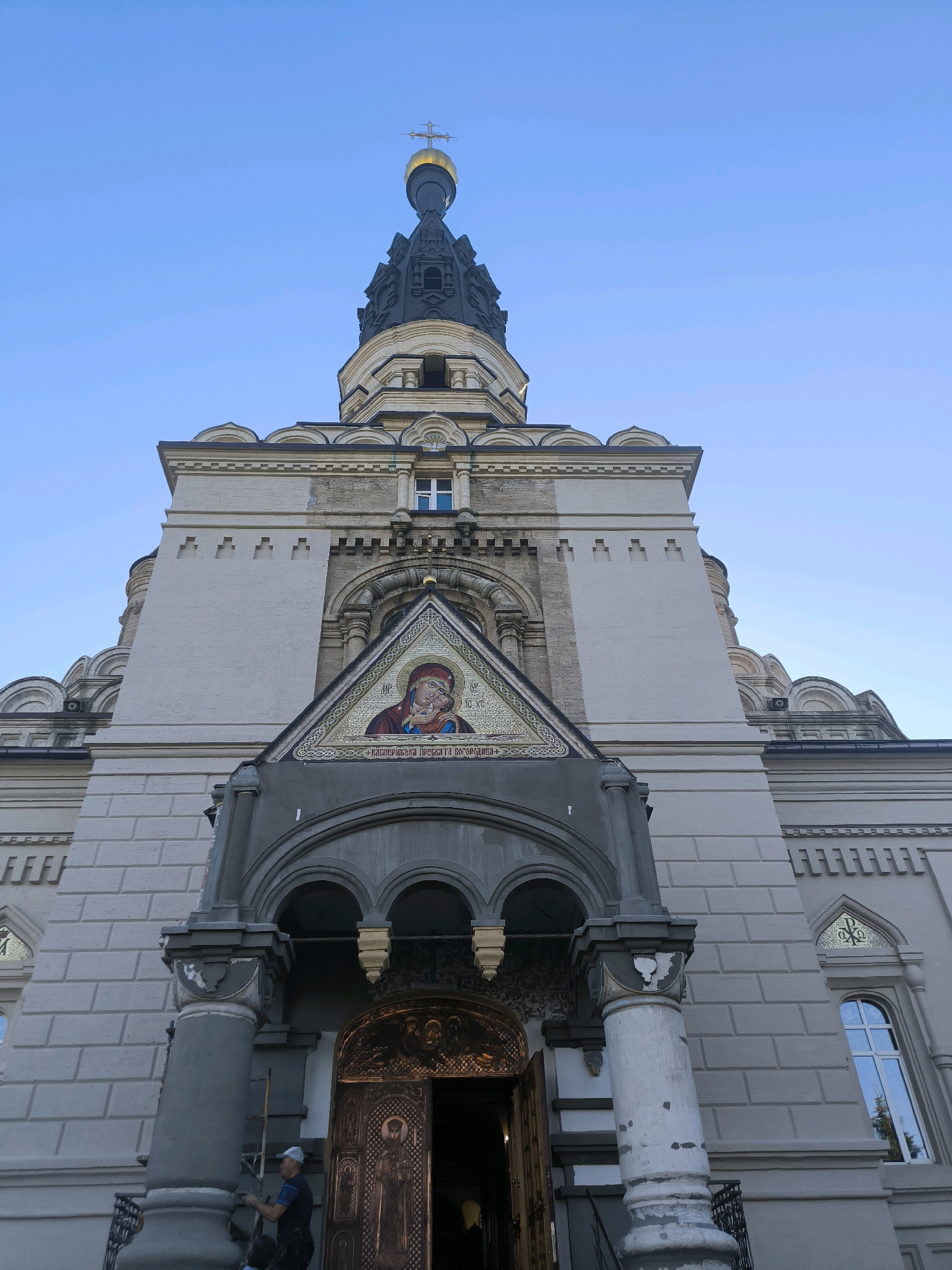 The image shows the façade of a church in Mykolaiv, Ukraine, under a clear blue sky. The building is constructed of light-colored stone and features a large, ornate golden dome topped with a cross on a tall, dark steeple. The entrance is framed by an arched portico with decorative columns, and above it, a triangular mosaic depicts a religious figure. The main body of the church has a textured stone facade with arched windows. The lighting suggests it is late afternoon or early evening. A person wearing a white cap and dark clothing is visible on the left side of the entrance, appearing to be working on the building.