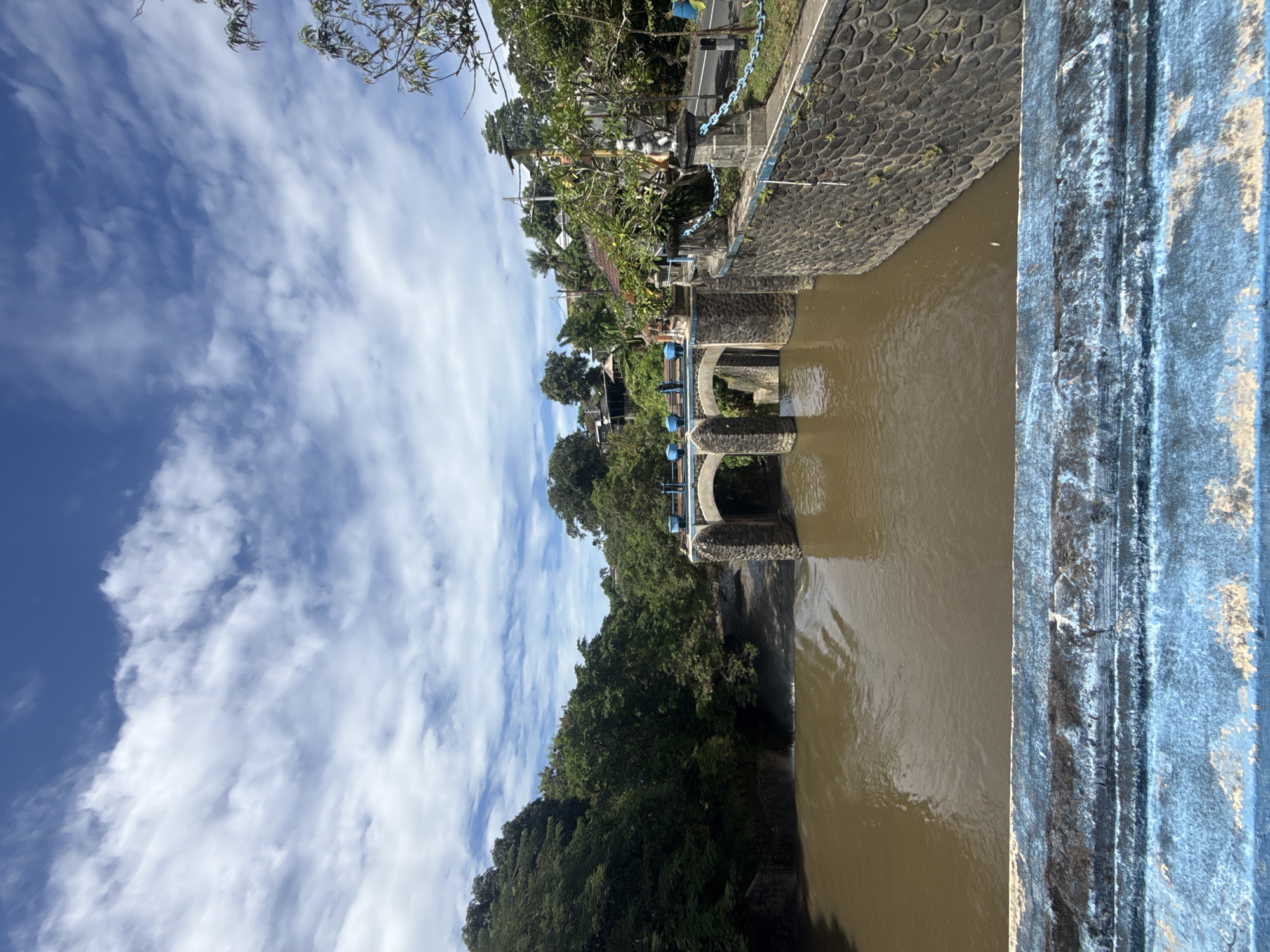 The image depicts a section of the Tukad Bindu river in Denpasar, Indonesia. The river is characterized by brown, turbid water flowing between stone banks and structures. A weir or dam is visible, constructed of stone arches and a blue-painted concrete structure at the right edge of the frame. A stone wall lined with a blue chain extends along the bank above the weir. Lush green vegetation covers the opposite bank, extending up a hillside. The sky is visible above, featuring a mix of blue sky and scattered white clouds.