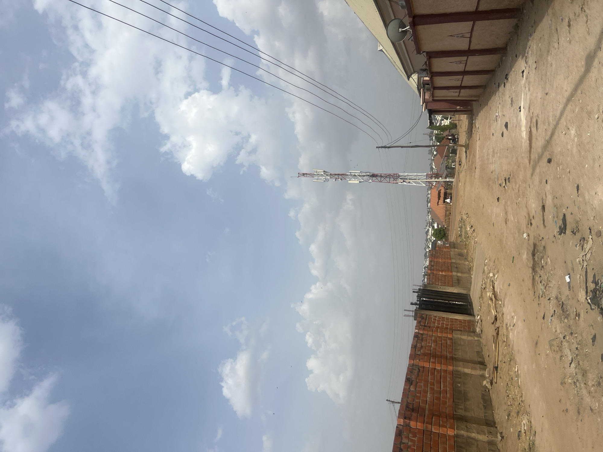 The image shows an outdoor scene in Minna, Nigeria. The sky is cloudy with a mix of white and gray clouds, with some power lines stretching across the upper portion of the frame. A communication tower, painted white and red, stands in the distance. A brick structure, possibly a wall or a building under construction, is situated to the right. The ground appears dusty and littered. Part of a building with an overhanging roof is visible along the right edge. In the background, a cluster of buildings indicates an urban or suburban environment. No people are directly visible in the foreground, but a blurry object appears in the background which is presumably a building with the rooftop of an orange hue.
