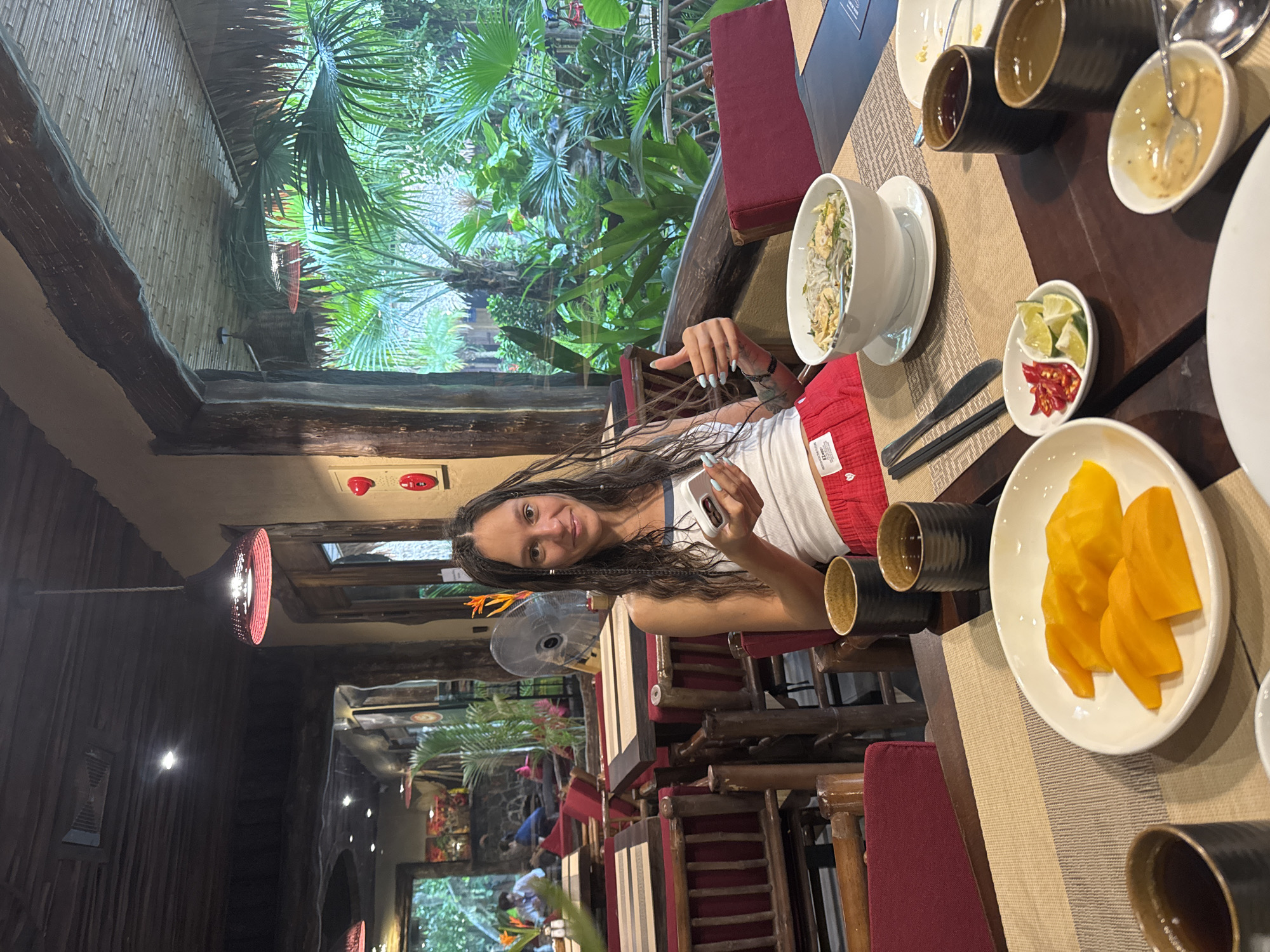 A woman with long, braided dark hair sits at a table in an open-air restaurant in Nam Hoa Lư Ward, Vietnam. She is wearing a white tank top and red shorts. She is holding a smartphone in her right hand and giving a thumbs up with her left hand. In front of her are a bowl of pho, a plate of sliced mango, and several cups of tea. The restaurant appears to be made of wood and bamboo, and lush green foliage is visible through an open doorway in the background. The lighting is warm and inviting.