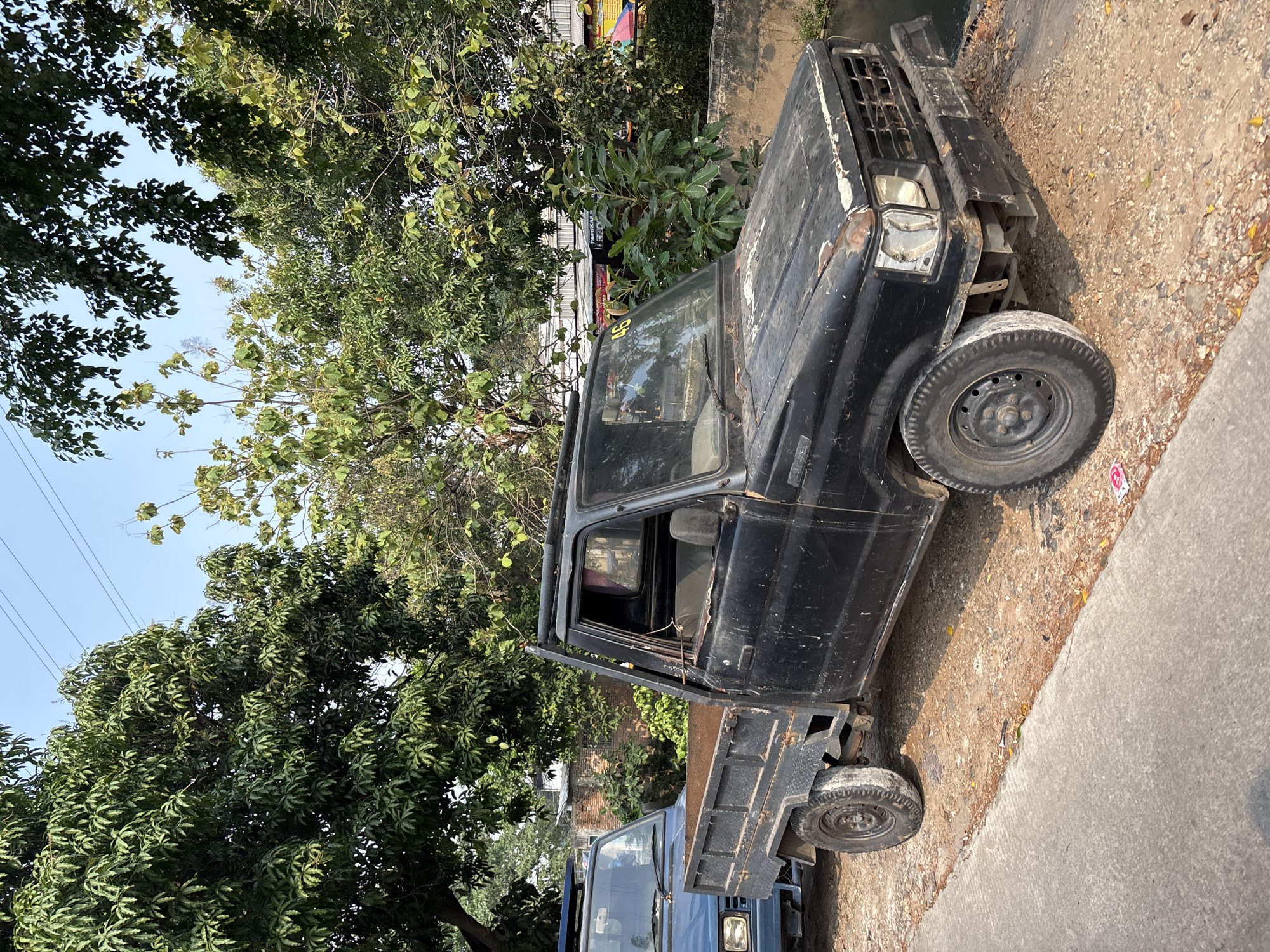 In Bekasi, Indonesia, a black pickup truck sits parked on a dirt and gravel shoulder next to a concrete pathway. The truck is older, showing signs of wear and tear, with a rusted bed and a dented front bumper. The driver's side door is open, revealing a worn interior. Another blue pickup truck is parked in front of it, partially visible. The scene is framed by lush green trees and a clear blue sky, suggesting a warm, sunny day. In the background, several buildings with tin roofs and brick walls are visible, as well as some colorful awnings that may indicate a market or food stalls. A narrow body of water runs along the right side of the image.