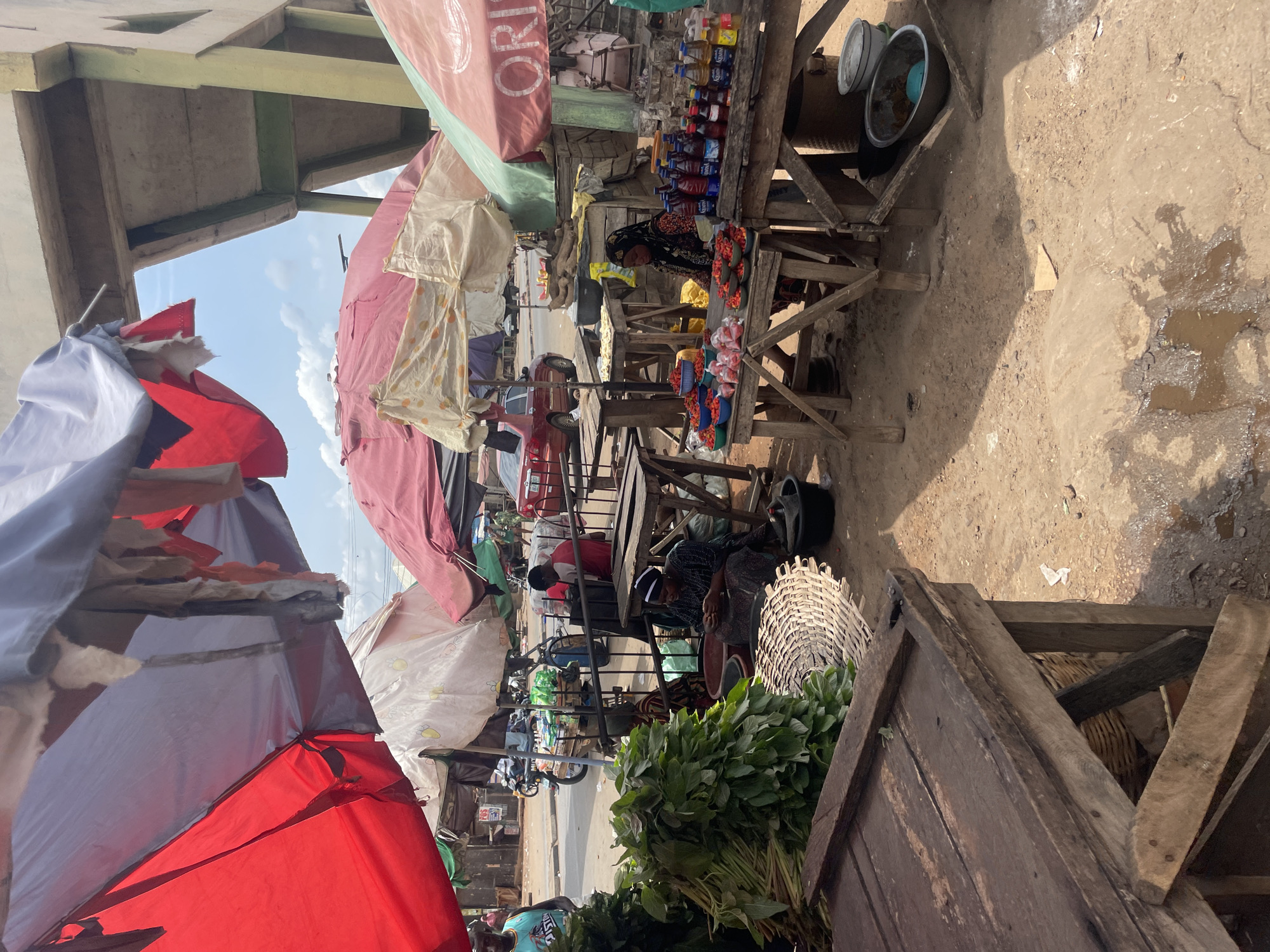 The image depicts a market scene in Lokoja, Nigeria. Stalls are set up with wooden tables displaying goods under makeshift shelters. People are present, some selling items like beverages, snacks, footwear, and vegetables. The ground is dirt and shows signs of wear. The shelters are made of colorful tarps and umbrellas.