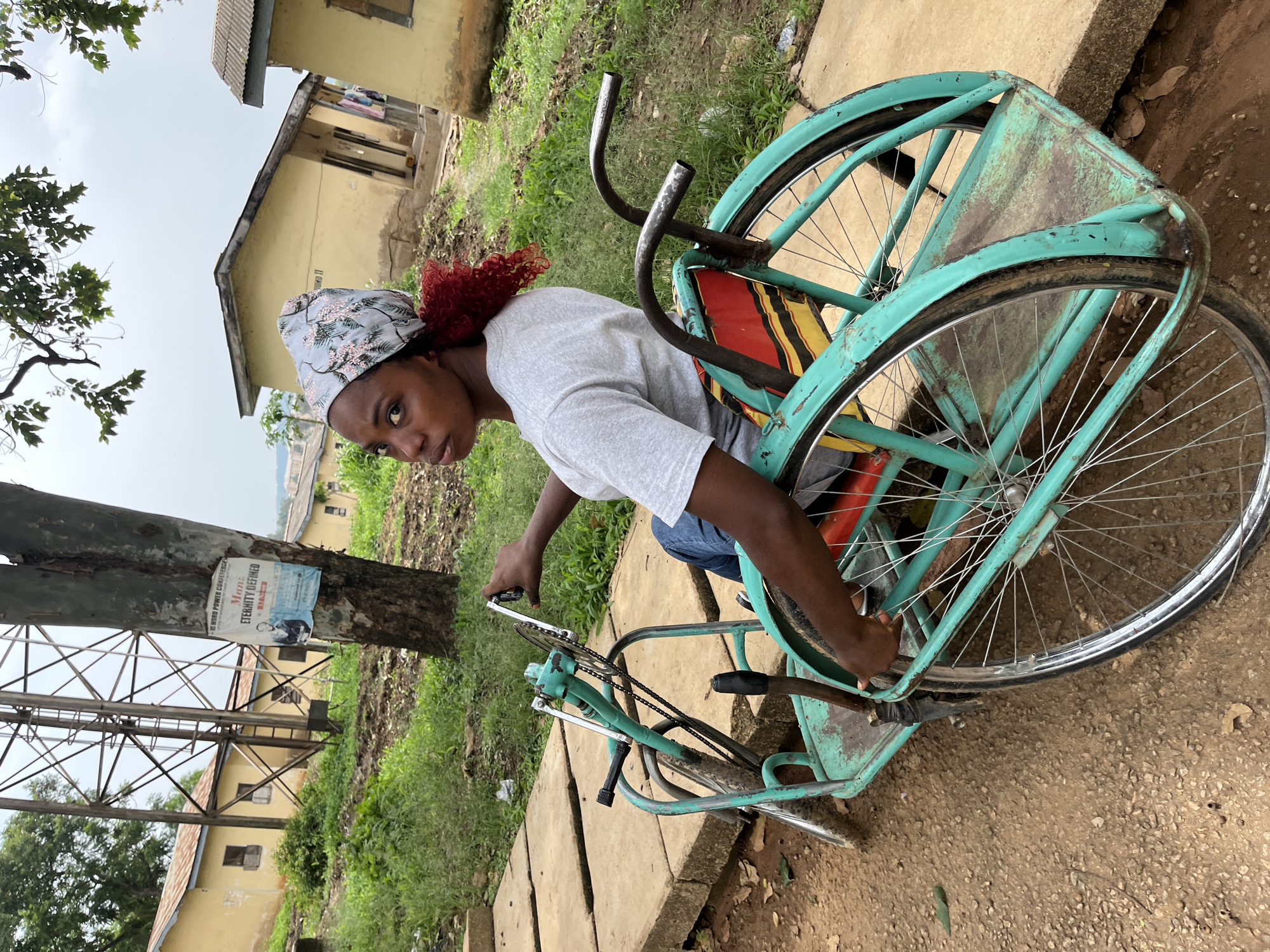 A young woman with bright red curly hair and a floral headscarf sits in a teal-colored, three-wheeled wheelchair. She is wearing a light grey t-shirt and blue jeans, and is looking back over her shoulder at the camera with a serious expression. Her left hand is on the wheelchair's handlebars, and her right hand is also resting on the wheelchair. The wheelchair appears to be made of metal and has large spoked wheels.
The scene is set outdoors, with a dirt and gravel path in the foreground. Behind the wheelchair, there are several yellow buildings with corrugated metal roofs, some of which appear to be in disrepair. A tall metal tower or structure is visible in the background, and there is lush green vegetation growing around the buildings and the path. A poster is attached to a tree in the middle ground, featuring text and a picture of a man. The sky is overcast and appears to be a light grey. The image was taken in the city of Rafin Par, Nigeria.