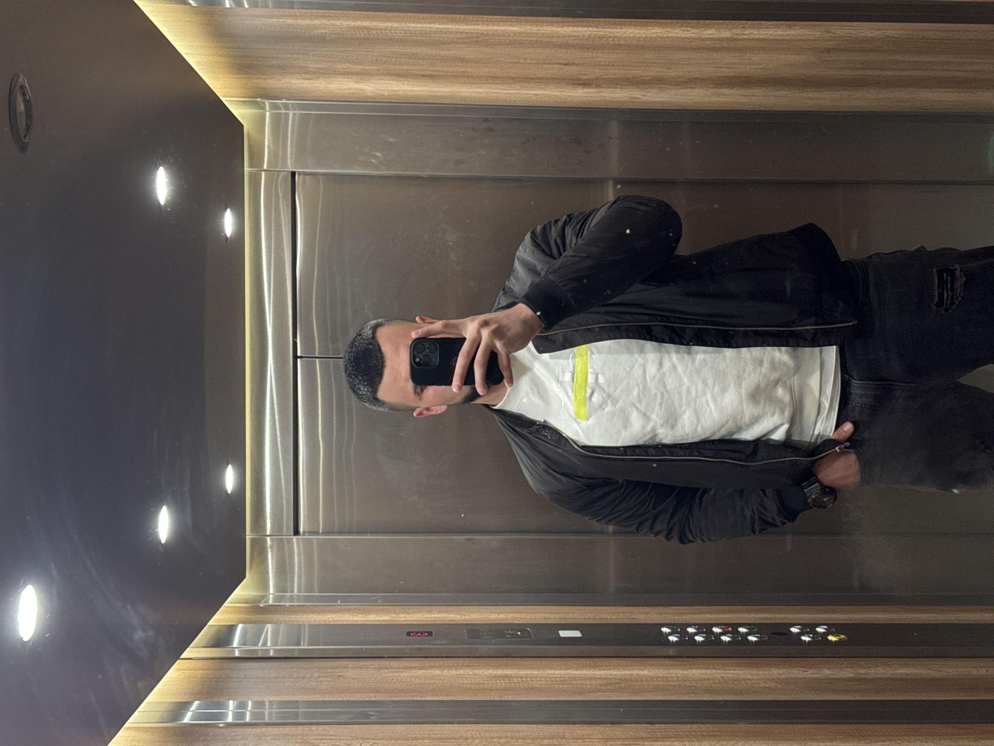 A man takes a selfie in an elevator. The elevator has gray panels, a mirrored wall with embedded lights, and a wood panel near the elevator control buttons. The man is wearing a black jacket, a white t-shirt with a yellow stripe, and black pants with a hole in the knee. He is holding a black phone in his left hand to take the picture. The location is unclear, but the city is Sofia, Bulgaria.