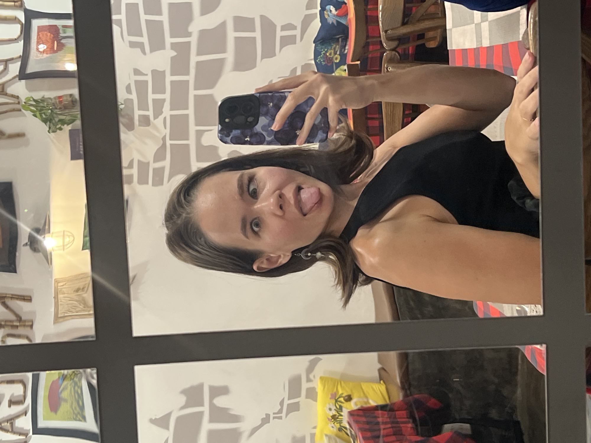 A woman takes a selfie in a mirror in Nha Trang Ward, Vietnam. The woman is sticking out her tongue and holding a phone with a floral case. The mirror reflects a room with artwork, a decorative wall, pillows, and chairs.