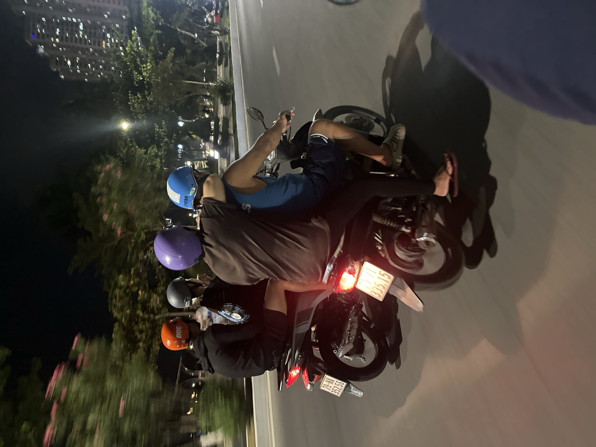 Four people are riding a motorcycle in Bac Nha Trang Ward, Vietnam, at night. The passengers are wearing helmets of different colors: orange, gray, purple, and blue. The motorcycle's license plates read "79-Y1 335.15" and "79-NA 252.56". High-rise buildings and trees line the road. The lighting suggests it is after dark.