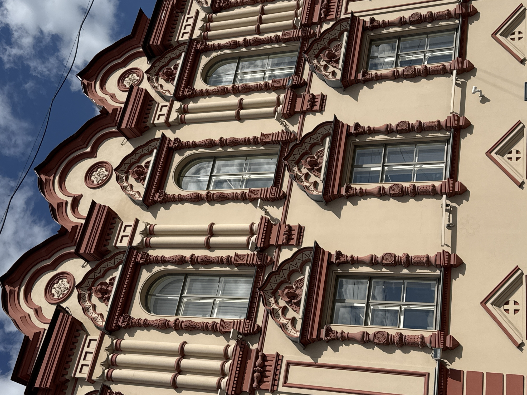 The image shows the facade of a building in Saint Petersburg, Russia. The architectural style is ornate, with a light beige base color and prominent dark red detailing. There are three visible levels, each featuring multiple windows. Some windows are arched, while others are rectangular. The windows are surrounded by decorative molding, which includes geometric patterns, stylized floral motifs, and small sculptures. Between the windows are alternating designs of horizontal cylindrical bands and decorative panels. The facade is oriented at an angle to the camera, and part of the sky with scattered clouds is visible in the upper left corner. There is also what appears to be an electrical wire running down from the top left corner. A security camera can also be seen mounted on the wall.