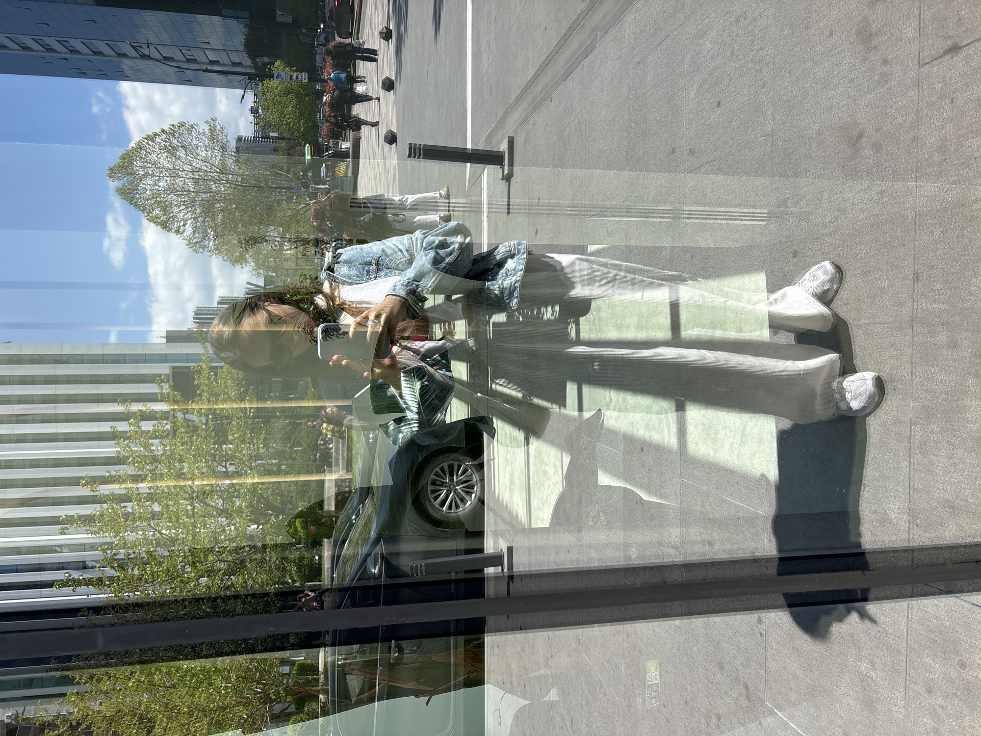 A person is captured taking a full-body selfie in the reflective glass facade of a modern building, likely in an urban setting. The image is taken outdoors on a bright, sunny day.
The primary subject is a young person, seemingly female, with long brown hair partially covering her face, holding a silver-colored smartphone to take the photo. She is dressed in a light-washed denim jacket over a white top, paired with wide-leg light-colored pants and white athletic sneakers. Her shadow is cast long on the light gray tiled pavement in front of her, indicating the sun is relatively low in the sky, suggesting either morning or late afternoon.
The reflection in the glass also shows the surrounding urban environment. Behind the person, a tall building with horizontal window lines is visible. To the left, a large, leafy tree with a rounded canopy stands prominently against a bright blue sky with scattered white clouds. Further in the background, more buildings and greenery are reflected. Parts of a dark-colored car, specifically its wheel, are also visible through the reflection. The ground area outside the building features a clean, light gray tiled pavement with several dark, cylindrical bollards or posts.
The reflections create a layered visual effect, superimposing the immediate foreground with the distant background. On the pavement, near the lower right corner, a small, faint yellow-green square marking with partially legible text, possibly "Tbil..." and "AL", is visible. The bright conditions and clear reflections suggest good weather. The location is contextualized as Tbilisi, Georgia.