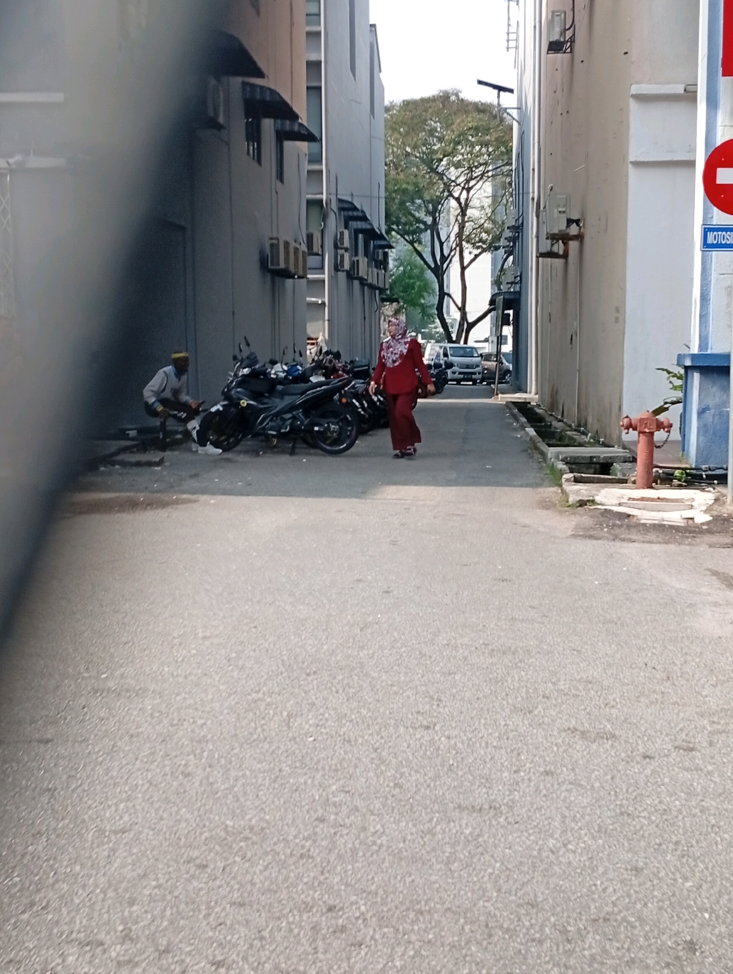 The image depicts an outdoor scene, likely in a city, with a narrow asphalt pathway flanked by buildings on both sides. In the foreground, the pathway is empty, but further down, a group of motorcycles are parked. To the left, a person in a light-colored jacket and dark pants, wearing a yellow head covering, is seated on a low stool next to a motorcycle. To the right, a person wearing a dark red hijab and matching outfit is walking away from the viewer, also near parked motorcycles. The buildings appear to be multi-story structures with visible air conditioning units. A fire hydrant is visible on the right side of the pathway. Trees are visible in the background, suggesting an urban environment with some greenery. A sign with a red circle and a white horizontal line is also present on the right. The lighting indicates it is daytime.