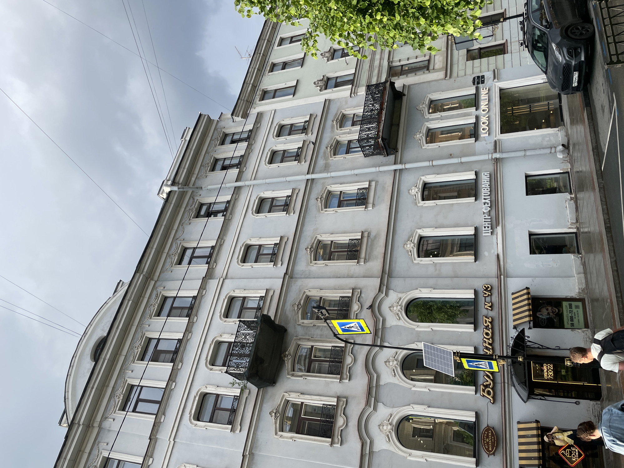 The photograph captures the detailed facade of a multi-story building on an overcast day, consistent with an urban setting in Saint Petersburg, Russia. The building is painted a light grey-blue and features ornate architectural details, including decorative window frames, some with arched tops, and several small, black wrought-iron balconies. Utility lines are visible against the cloudy sky in the upper left, while green tree leaves frame the upper right corner.
At street level, the pavement appears damp, suggesting recent rain. A dark grey car, bearing the Russian license plate "P184MO 198", is parked at the curb. The ground floor houses several businesses, identified by visible signage. "LOOK ONLINE" is prominently displayed, along with "ЦЕНТР ФЕХТОВАНИЯ" (Fencing Center). Further along, a large gold sign reads "Булочная N 43" (Bakery No. 43), accompanied by smaller signs for "Пекарня Вольчека" (Volchek's Bakery) and "КОФЕ" (Coffee). Operating hours are indicated by an "ОТКРЫТО 7:30-22" (Open 7:30-22) sign. Striped awnings are seen above one of the shop entrances.
A yellow pedestrian crossing sign, featuring a blue figure, is mounted on a pole that also supports a small solar panel. Two men are visible on the sidewalk; one in a white t-shirt with a dark backpack, and another in a blue shirt. The image portrays a typical daytime street scene, with commercial activity and passersby, under a cloudy sky.