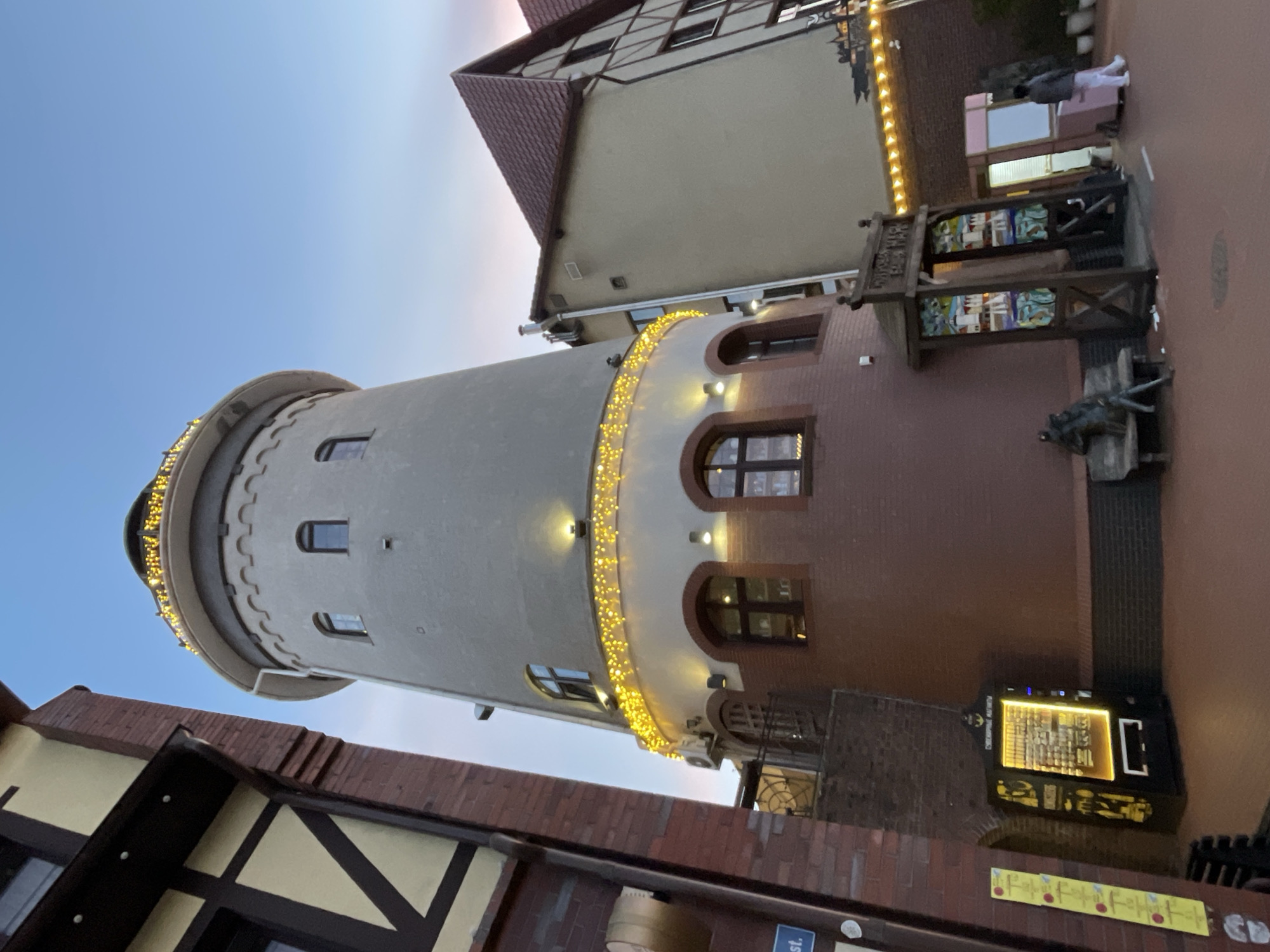 The image shows a tall, cylindrical tower in Kaliningrad, Russia. The tower is mostly light gray, with a section near the base made of red brick. Arched windows are present at the base and higher up on the red brick section. The top of the tower is adorned with small, rounded battlements. A string of yellow lights is wrapped around the tower at two different levels. Adjacent to the tower are buildings with German-style architecture, including timber framing and tiled roofs. A sign with an intricate wooden frame is also visible, as is a modern electronic information kiosk on the brick section of the tower. The sky is a gradient of blue and pink, indicating either dusk or dawn.