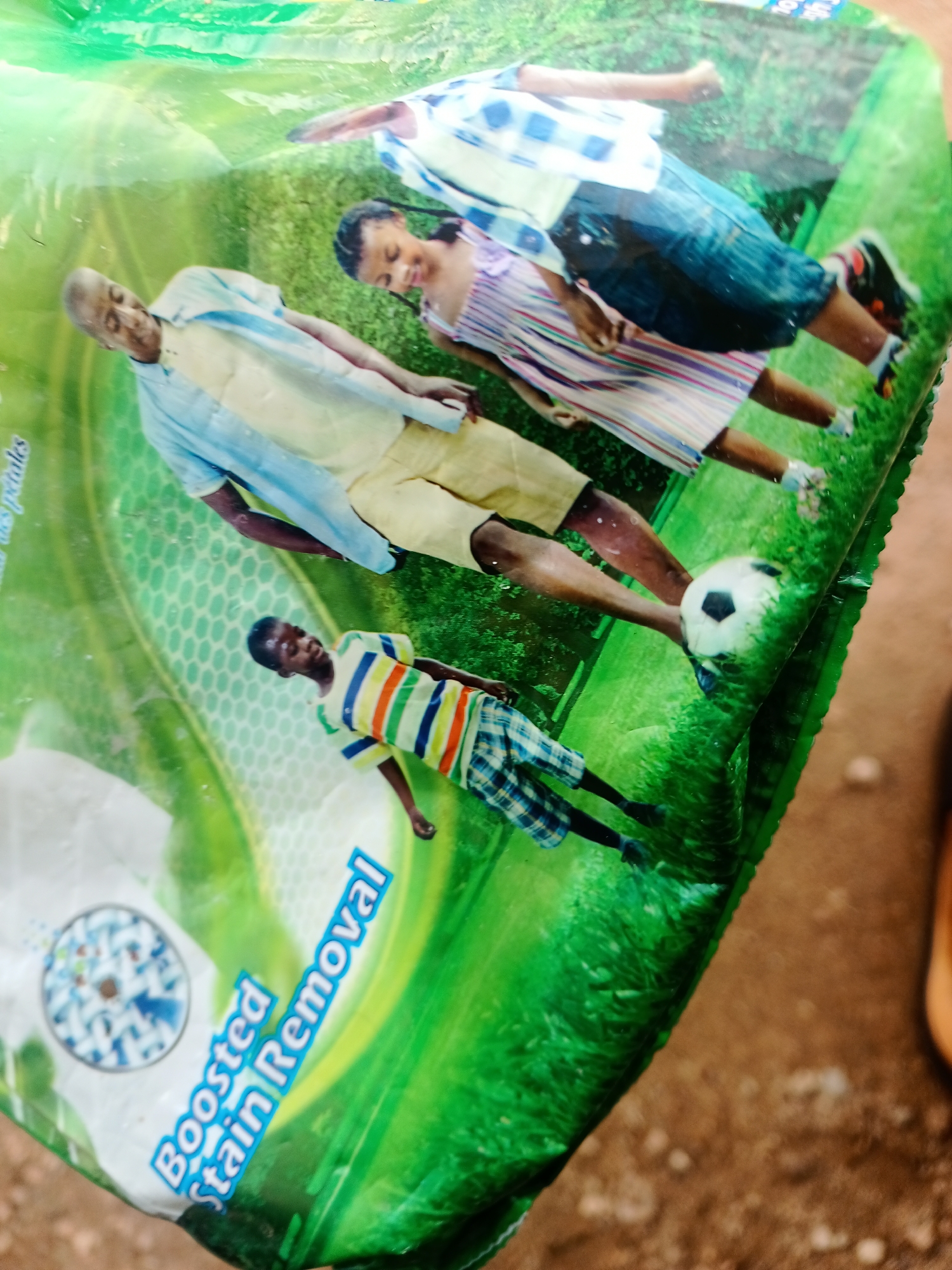 The image shows a close-up of a detergent packaging with a family graphic. The family appears to be of African descent and includes a man, a woman, and two children. They are depicted in an outdoor setting, possibly a park or garden, with green grass and a soccer ball. The man is wearing a light blue shirt and yellow shorts, the woman is in a striped dress, one child is in a plaid shirt and shorts, and the other is in a striped t-shirt and plaid shorts. The packaging also features the text "Boosted Stain Removal". The image is taken at an angle, suggesting it might be a product photo or a consumer showcasing the product. There is no visible indication of the specific location in Jalingo, Nigeria, other than the product packaging itself, which might be popular in that region.
