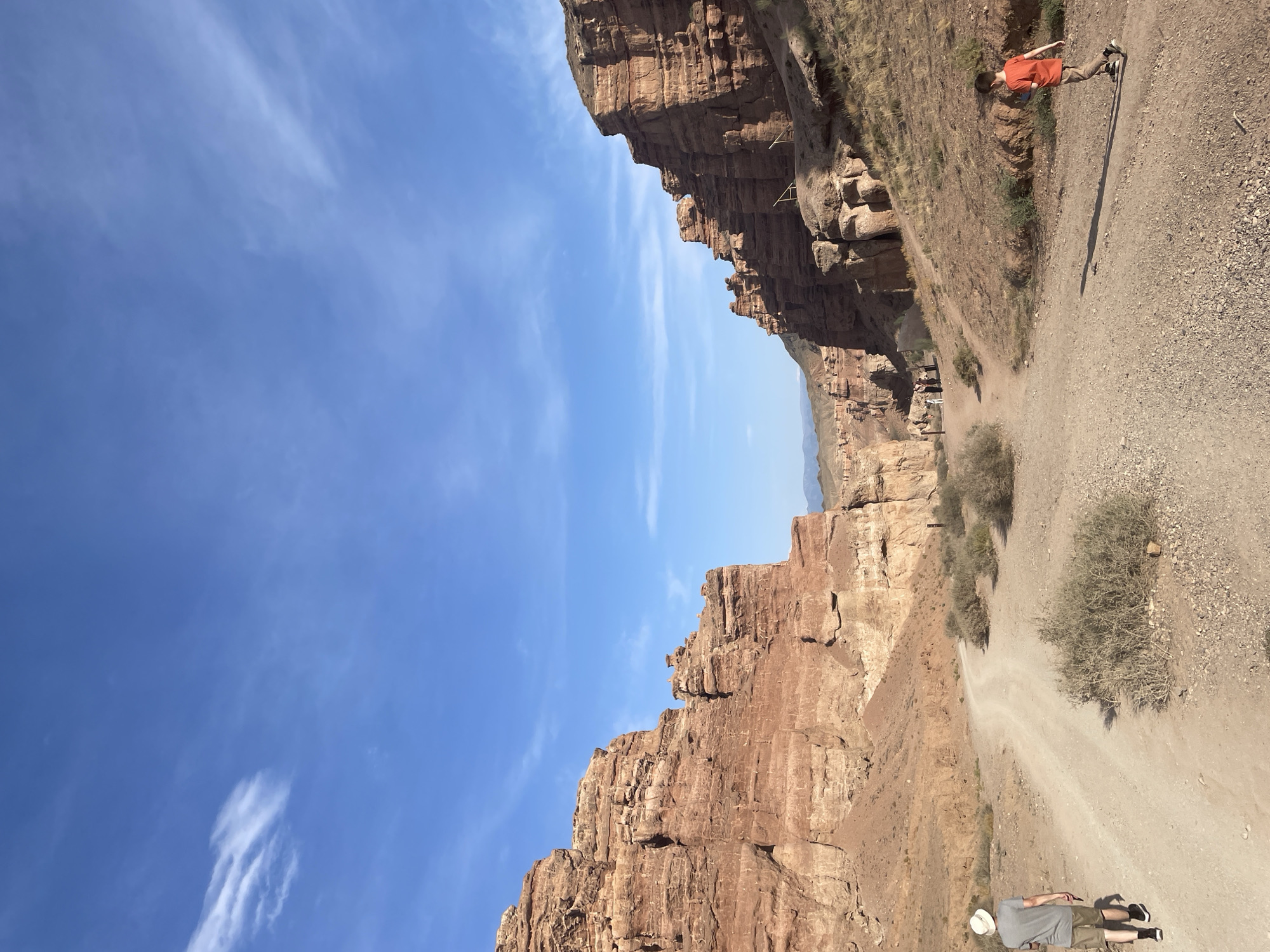 A wide shot captures the Charyn Canyon in Tamerlik, Kazakhstan, under a clear blue sky dotted with wispy clouds. The canyon walls, composed of reddish-brown sedimentary rock, rise steeply on either side of a dusty, unpaved path that winds through the center of the frame. Sparse, dry vegetation, including scrubby bushes and grasses, grows on the slopes.
In the foreground, two individuals are walking on the path. On the left, a person wearing a light-colored hat, a grey t-shirt, and shorts walks away from the camera. On the right, a young person in an orange t-shirt and tan pants walks towards the right side of the frame, casting a shadow on the path.
Further into the canyon, a group of people are visible, appearing to be exploring the area. The path continues to recede into the distance, framed by the imposing canyon walls. The overall scene depicts a natural landscape of geological formations with human presence engaging in outdoor activity.
