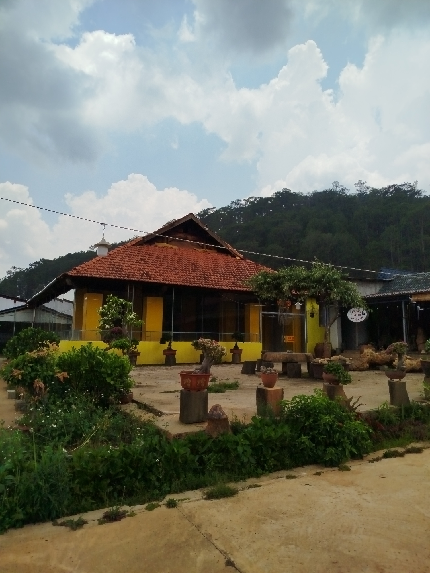 The image shows a yellow-painted building with a red tile roof, located in Hiep Thanh, Vietnam. The building features large glass windows and is surrounded by a garden area with potted plants and greenery. A sign with Vietnamese script is visible near the entrance of the building. The surrounding environment includes a hillside covered with trees, and a concrete area in the foreground. The sky is cloudy.