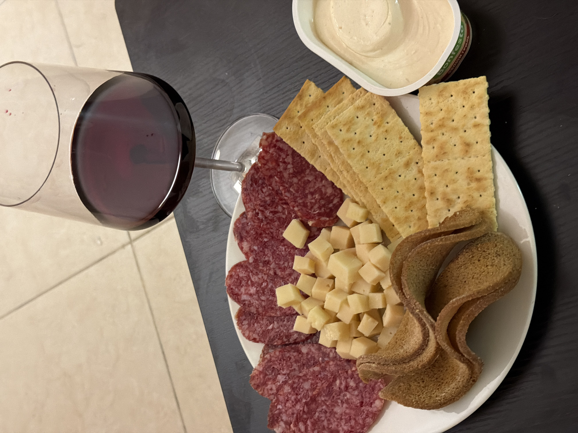 A red wine glass sits beside a plate of crackers, cubed cheese, salami, and brown bread. A container of white dip also sits nearby. The scene appears to be indoors, possibly on a table. There is no evidence in the image to suggest the location is Tel-Aviv, Israel.