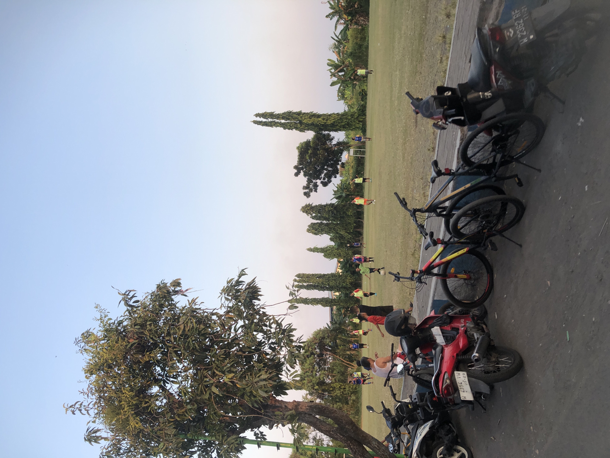 The image captures a grassy field in Tenggulunan, Indonesia, where a group of men are engaged in a soccer game under a clear, pale blue sky. The field is bordered by various trees and some banana trees. In the foreground, parked on a paved area, are several motorcycles and bicycles. One red motorcycle has a license plate reading "W 4352 VW" and "05-29." Another dark-colored motorcycle to its right has a license plate that is partially visible. Two bicycles are parked next to each other, with the one closer to the motorcycles featuring a black frame with yellow and red accents on the wheels. A man in a white tank top and red shorts sits on a motorcycle, facing away from the camera. The overall scene depicts a casual recreational activity in an outdoor, semi-urban setting.