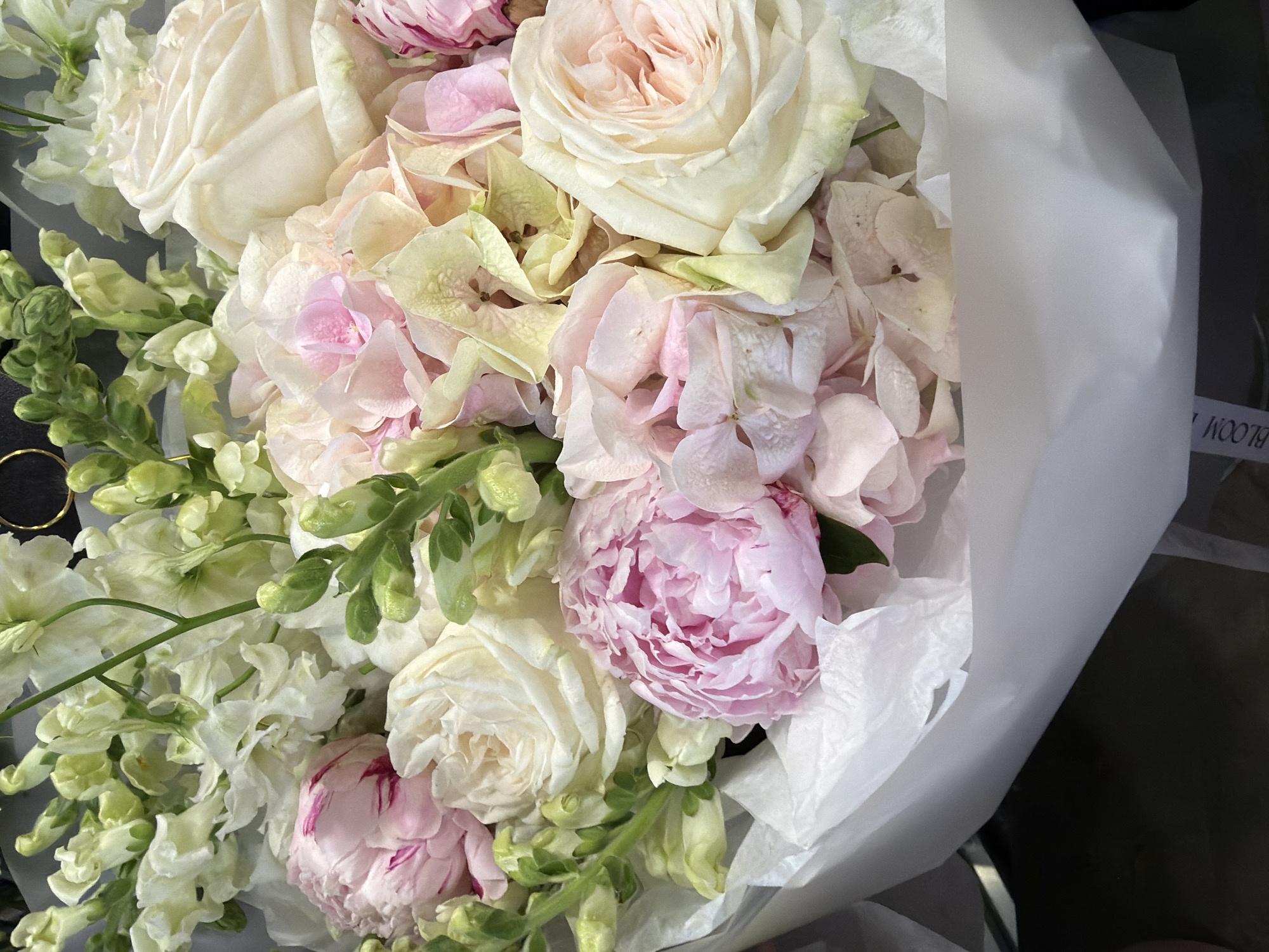 A close-up shot features a bouquet of flowers, primarily composed of light pink and cream-colored roses and peonies. The arrangement is wrapped in white tissue paper, which is slightly crumpled. The flowers are densely packed, creating a soft, textured appearance. The lighting suggests an indoor setting, possibly a florist shop or a home. The background is dark and out of focus, drawing attention to the floral display. A partial view of a dark surface, possibly a table or counter, is visible at the bottom of the frame.