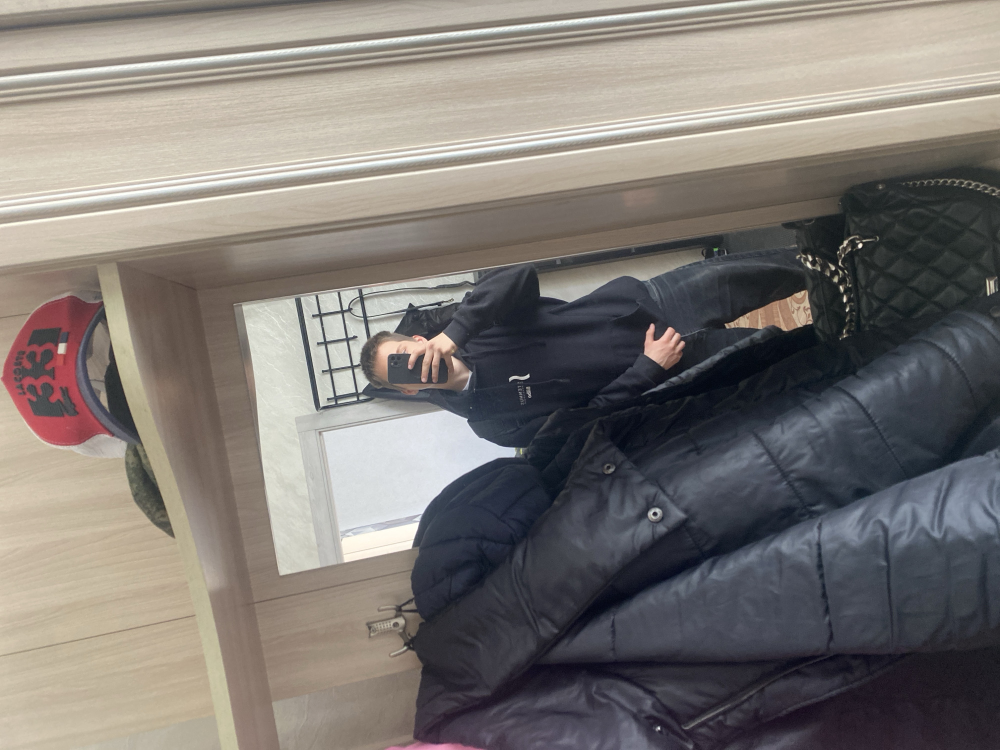 A person is taking a selfie in a mirror. The reflection shows the person wearing a black hoodie and jeans. A black purse and a dark, quilted jacket are near the mirror. A red and white hat and a metal rack are visible on the left. The background through the mirror includes a window. The location is Chaikovsky, Russia.