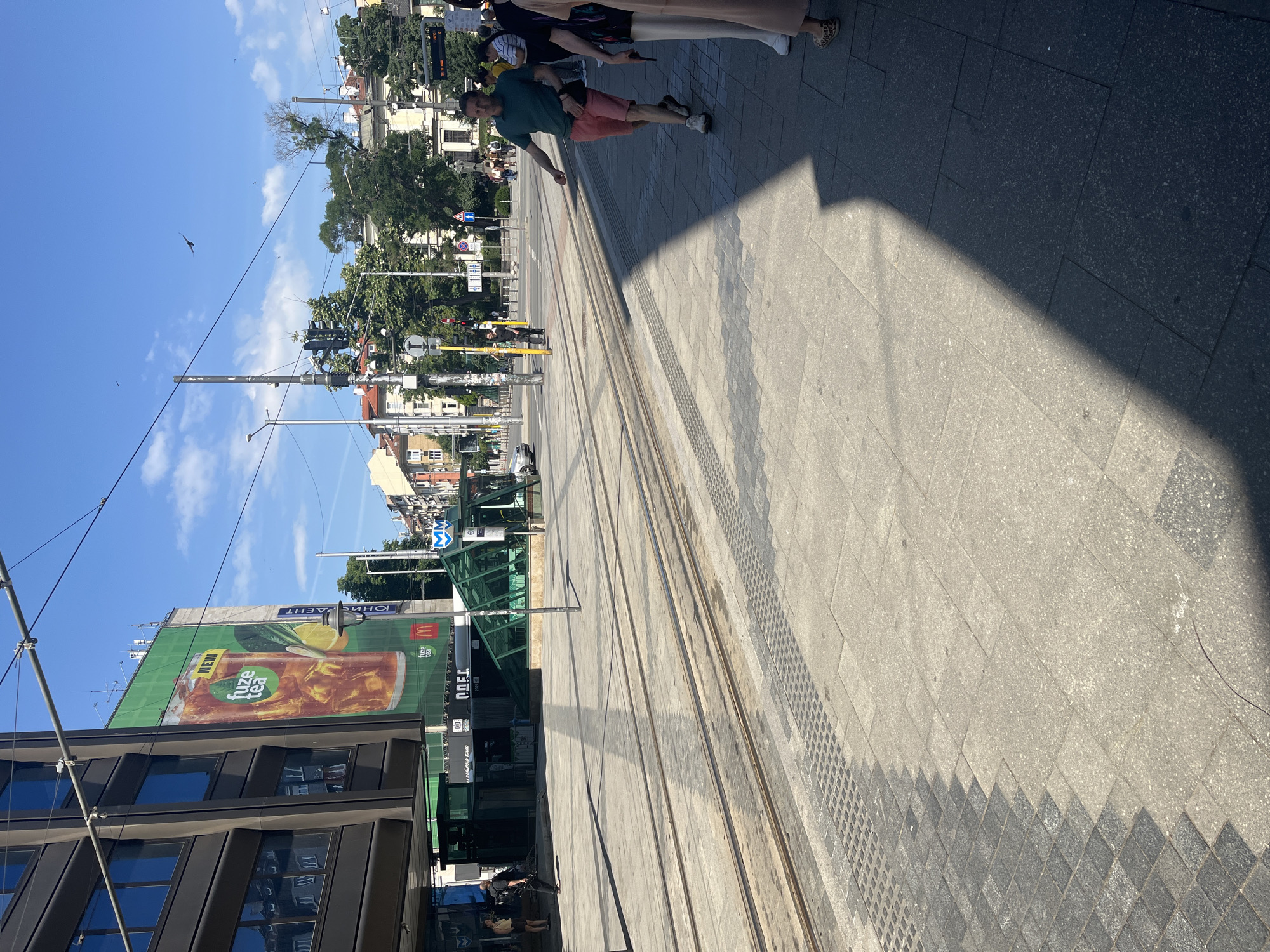 This is an eye-level, medium shot taken on a sunny day in Sofia, Bulgaria, likely near a tram station. The foreground shows paved ground with tram tracks running horizontally across the frame. The pavement is a mix of light gray and darker gray textured tiles. A sharp shadow of an unseen object bisects the lower right portion of the image, casting a dramatic contrast on the bright pavement.
In the mid-ground, to the left, a large building with dark tinted windows is visible. A massive green billboard advertising "Fuze Tea" with a large image of iced tea and a slice of lemon is plastered on the side of this building. Below the billboard, a green structure with a glass roof and entrance appears to be a tram or metro station entrance. Some text in Cyrillic is visible on the building and signage.
To the right of the tram tracks, a man in a green t-shirt and pink shorts is walking away from the camera. He has a dark fanny pack around his waist. Further to the right, the partial figures of at least two other people are visible, one wearing a light-colored skirt or dress and the other in a patterned garment.
The background reveals more of the city's architecture with multi-story buildings and trees. Traffic lights and street signs are also present, indicating an urban intersection. The sky is a clear blue with scattered white clouds, and a bird is captured in flight high above. The overall scene depicts a bustling urban environment on a bright day.