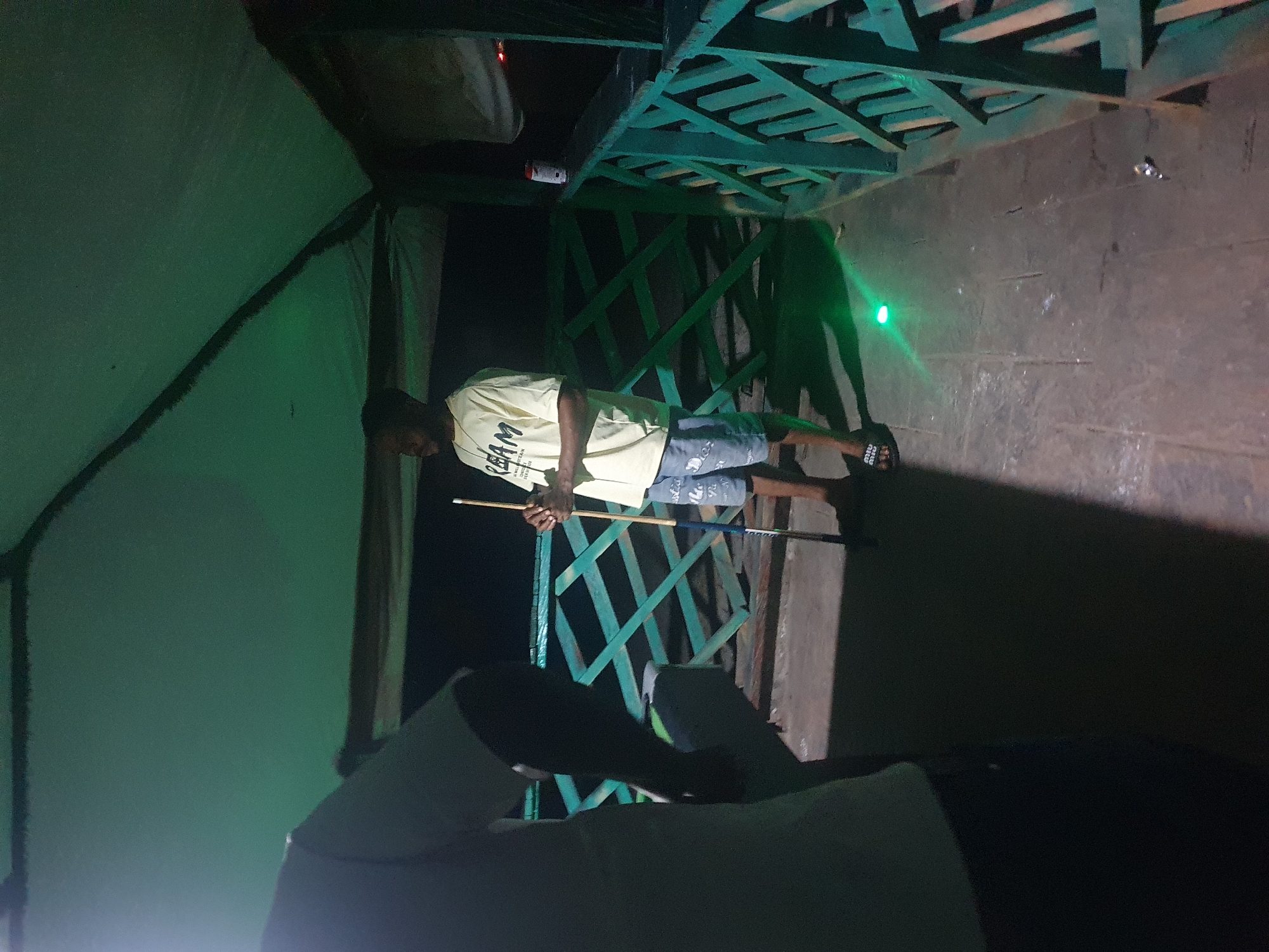 The image depicts a night scene, likely an outdoor recreational area with a canopy overhead. The environment appears to be a waterfront or a resort area in Lokoja, Nigeria, evidenced by the tropical flora and the general ambiance suggestive of a warm climate. The lighting is dim, with green hues dominating the scene, possibly from decorative lights or ambient illumination from a nearby establishment.
In the foreground, a person is partially visible on the left, wearing a dark shirt and pants. Their back is to the camera, and they are standing in shadow, suggesting they are a participant or observer in the activity.
In the mid-ground, a man stands facing to the left, holding a pool cue. He is wearing a light-colored t-shirt with text that appears to read "REAM," and blue shorts. He is standing on a tiled or concrete surface. His posture suggests he is either playing pool or preparing to play. To his right, a green railing with a diamond pattern is visible, characteristic of outdoor structures in such settings.
In the background, there's a hint of dark foliage and possibly other structures, obscured by the low light conditions. A red and white can is visible on the railing to the right of the man with the cue, suggesting the presence of refreshments. A faint green light emanates from the ground in front of the man, creating a pool of light on the surface. The overall atmosphere is relaxed and informal, indicative of leisure activities.
