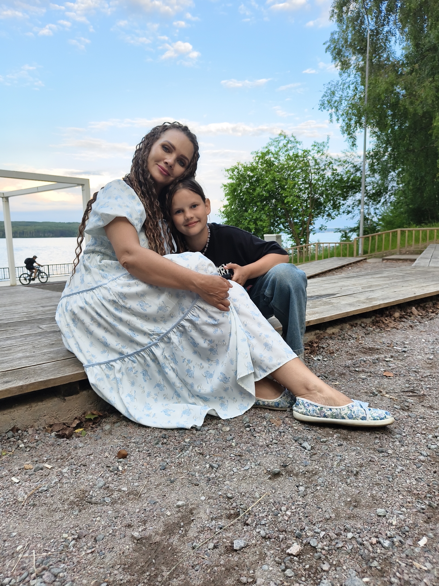 A woman and a child are sitting on a wooden deck near a body of water. The woman has long, braided, dark hair and is wearing a light blue dress with a floral pattern. She is smiling and has her arm around the child. The child has dark hair and is wearing a black t-shirt and blue jeans. The child is leaning against the woman and smiling. In the background, there is a lake, trees, and a clear sky with some clouds. A person is riding a bicycle in the distance. The setting appears to be a park or recreational area in the city of Chaikovsky, Russia.