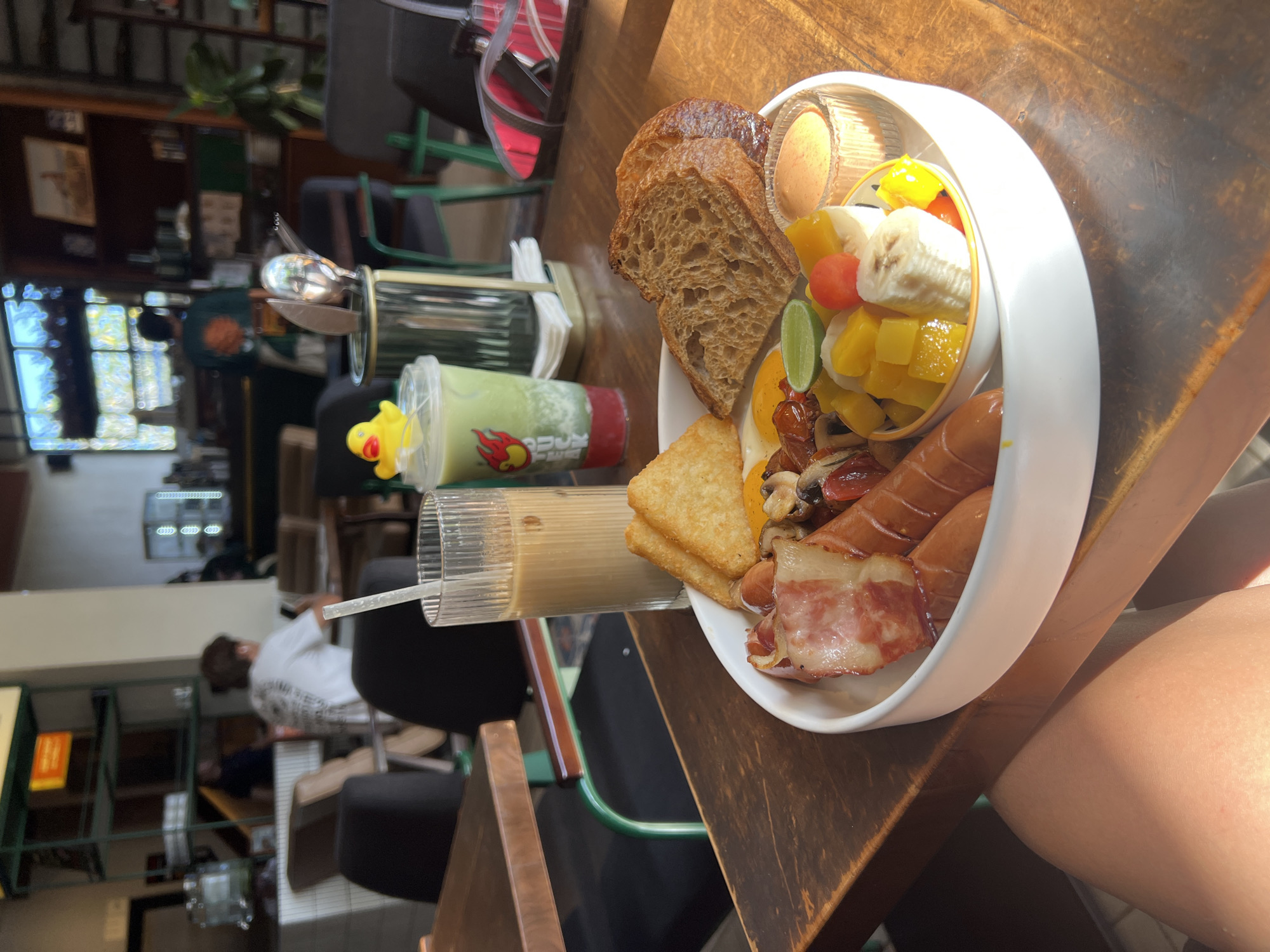 A view of breakfast on a wooden table is captured at a high angle in Kuta Selatan, Indonesia. The breakfast includes toast, a variety of fruits, including banana and mango, hash browns, sausage, and bacon, all neatly arranged in a large white bowl with a compartment for dipping sauce. Beside the bowl, a green smoothie and a glass of iced coffee sit on the table. A partial view of a person's leg is also visible. The background shows people sitting at tables in an indoor setting with glass windows, indicating a restaurant or cafe.