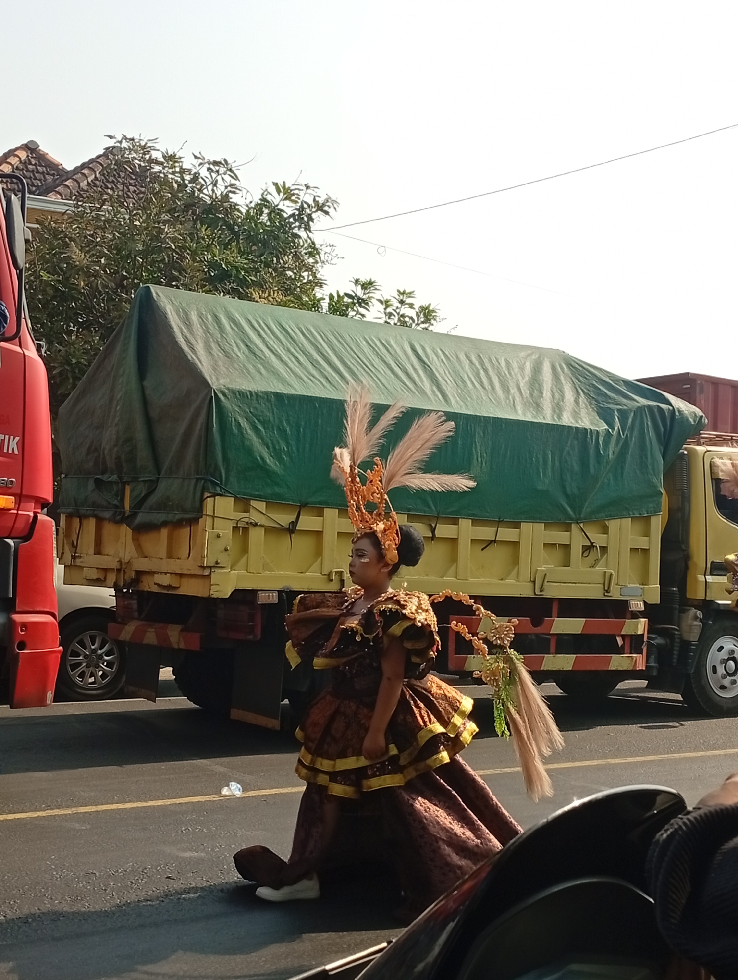 A woman in a large, ornate costume with feathers and gold embellishments walks along the side of a road in Sarang, Indonesia. The woman is wearing a brown and gold patterned dress with gold trim, and a elaborate headdress with feathers. She is wearing white sneakers. Behind her, a large yellow truck with a green tarp covering its cargo is parked on the side of the road. To the left of the truck, a red truck is partially visible. The road has yellow lines painted on it. Trees and a building with a tiled roof are visible in the background. The sky is clear and bright.