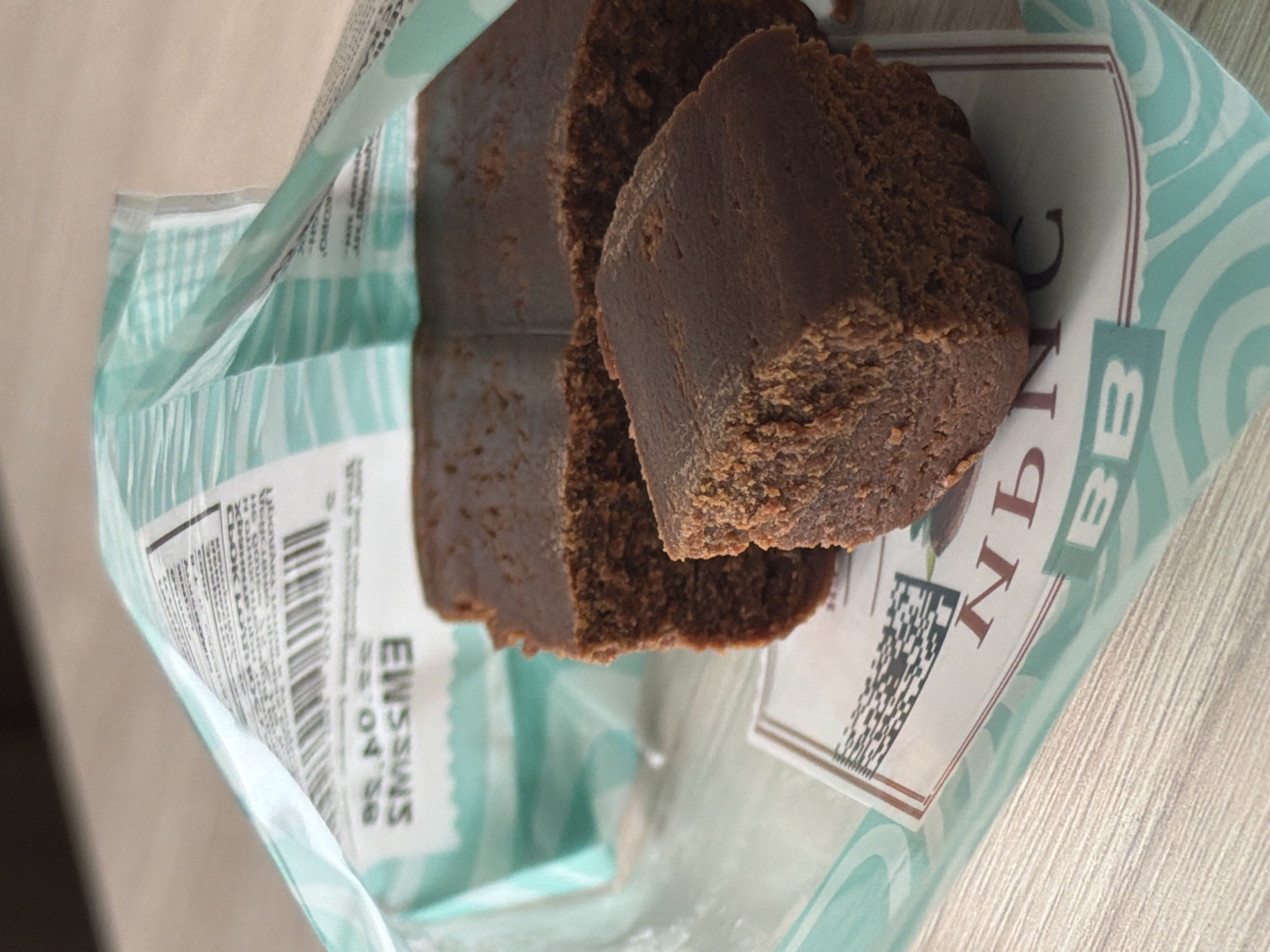 Here is a detailed description of the image:
The image shows pieces of what appears to be chocolate brownie or fudge in a light blue and white patterned wrapper or bag. The bag has printed text, including a nutrition information section and the partial word "EWSSWS." There is also a small label with text and a barcode, possibly part of the product packaging. The brownies are dark brown with a somewhat dense texture. The image is taken at a close angle, focusing on the product inside the open packaging. The surface beneath the packaging appears to be a light-colored, possibly wooden, surface. The style and branding indicate the product may be associated with "Saint Petersburg, Russia" as mentioned.
