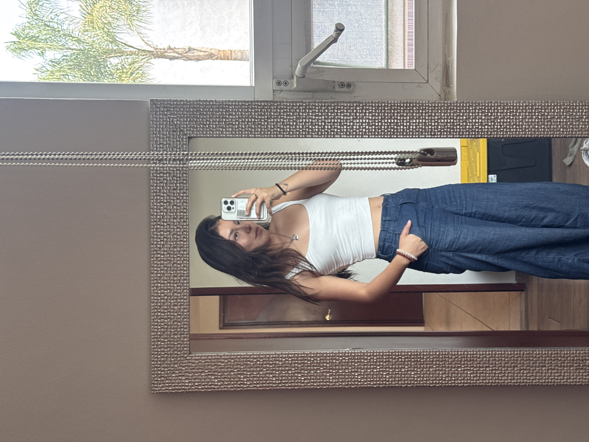 A woman stands in front of a large mirror in a room with a window. The mirror has a beaded frame. The woman is holding a smartphone up to take a picture of herself. She is wearing a white tank top and blue jeans. The window shows a tree and a cloudy sky. The mirror reflects a hallway with a partially visible door. The location appears to be inside a building in the city of Heroica Puebla de Zaragoza, Mexico.