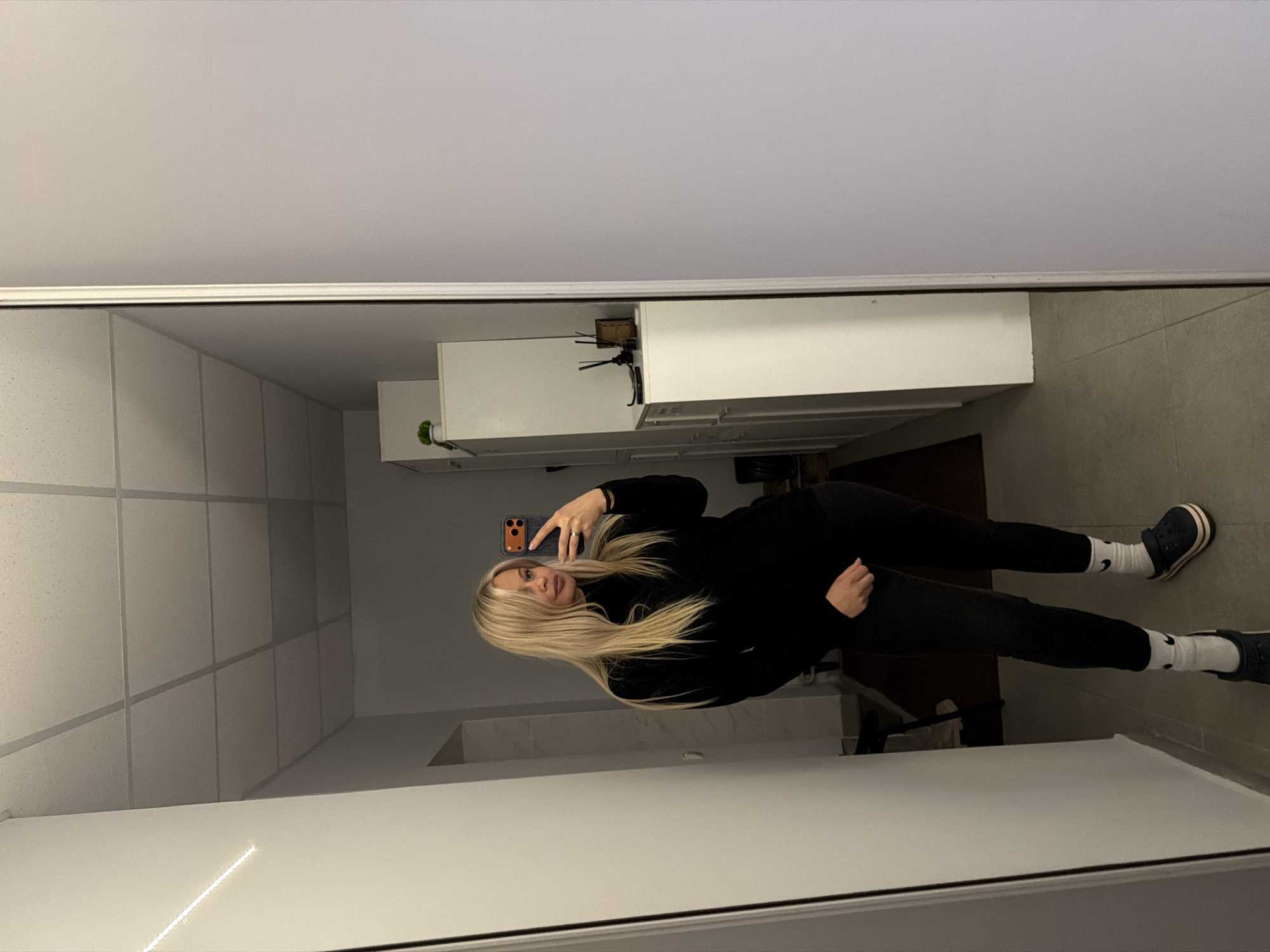 A woman is reflected in a mirror while taking a photo of herself with her phone. The woman is wearing a black top, black leggings, white socks, and blue shoes. Her hair is long and blonde. The background of the mirror's reflection shows a room with white walls, a white ceiling with a grid pattern, and white cabinets. A vacuum cleaning device is visible on top of one of the cabinets. The location is a generic interior; there is no definitive information to confirm it is in Sarapul, Russia.