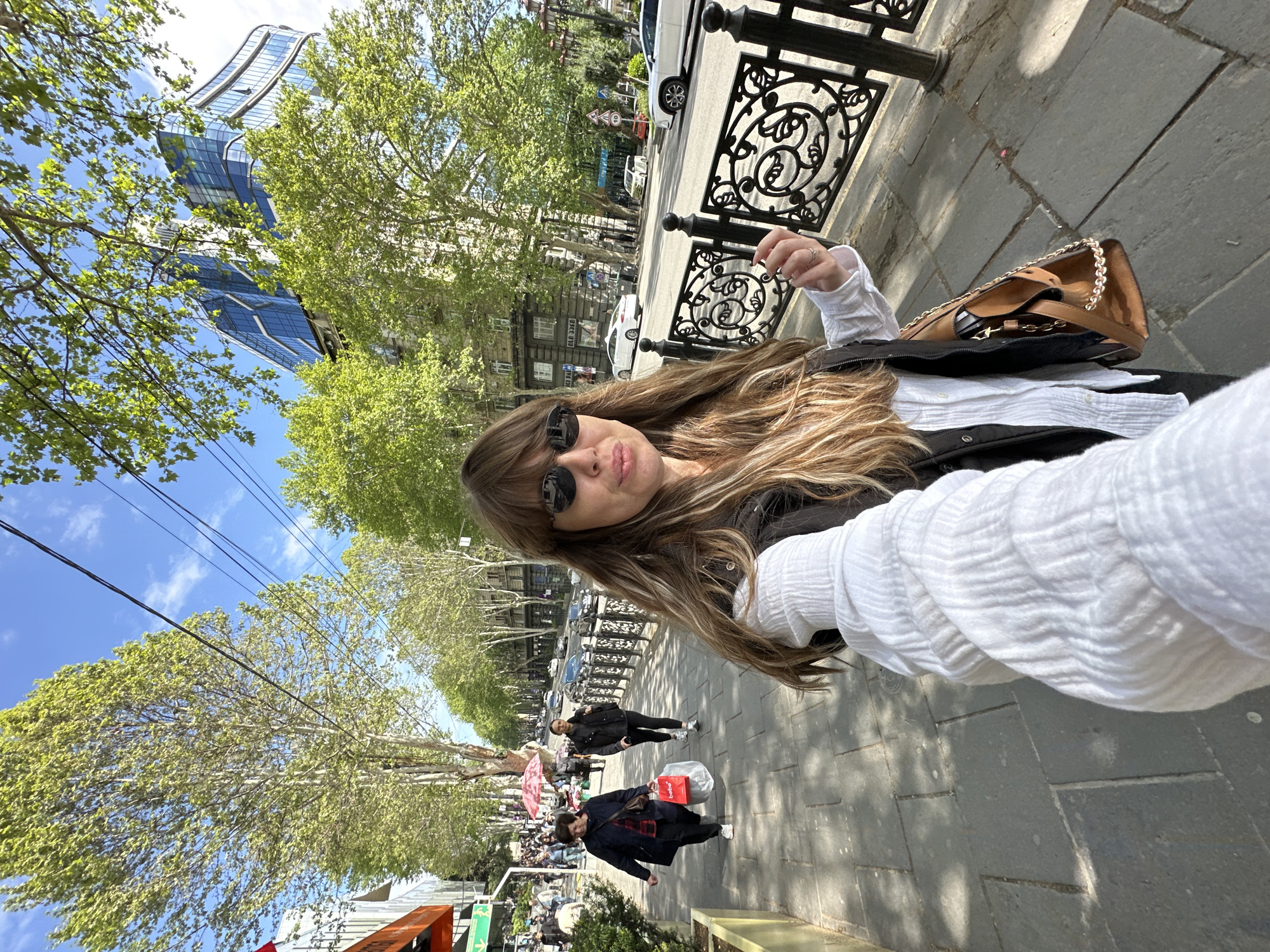 A woman is taking a selfie while standing on a sidewalk. She is wearing a white shirt, black jacket, and sunglasses, and is carrying a brown purse. Behind her is a street with trees lining the sidewalk. People are walking on the sidewalk and crossing the street. Cars are parked along the side of the street. A tall, modern glass building is visible in the background. The image was taken in Tbilisi, Georgia, on Rustaveli Avenue.