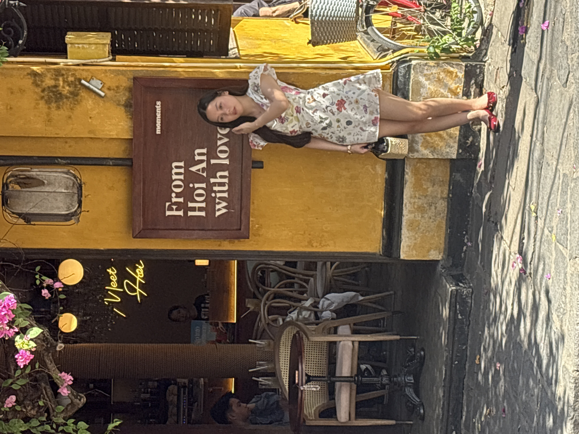 A young Vietnamese woman stands on a cobblestone street in front of a yellow building in Hoi An Ward, Vietnam. She is wearing a short, floral print dress and red ballet flats. In her right hand, she holds a straw handbag. Behind her, a neon sign in a window reads "Meet Hoi". A wooden sign on the building reads "From Hoi An with love". A bicycle with a basket is parked to the right of the woman. Inside the building, tables and chairs are visible, suggesting it is a cafe or restaurant. The ground is scattered with fallen pink flower petals. The sunlight casts dappled shadows on the street.