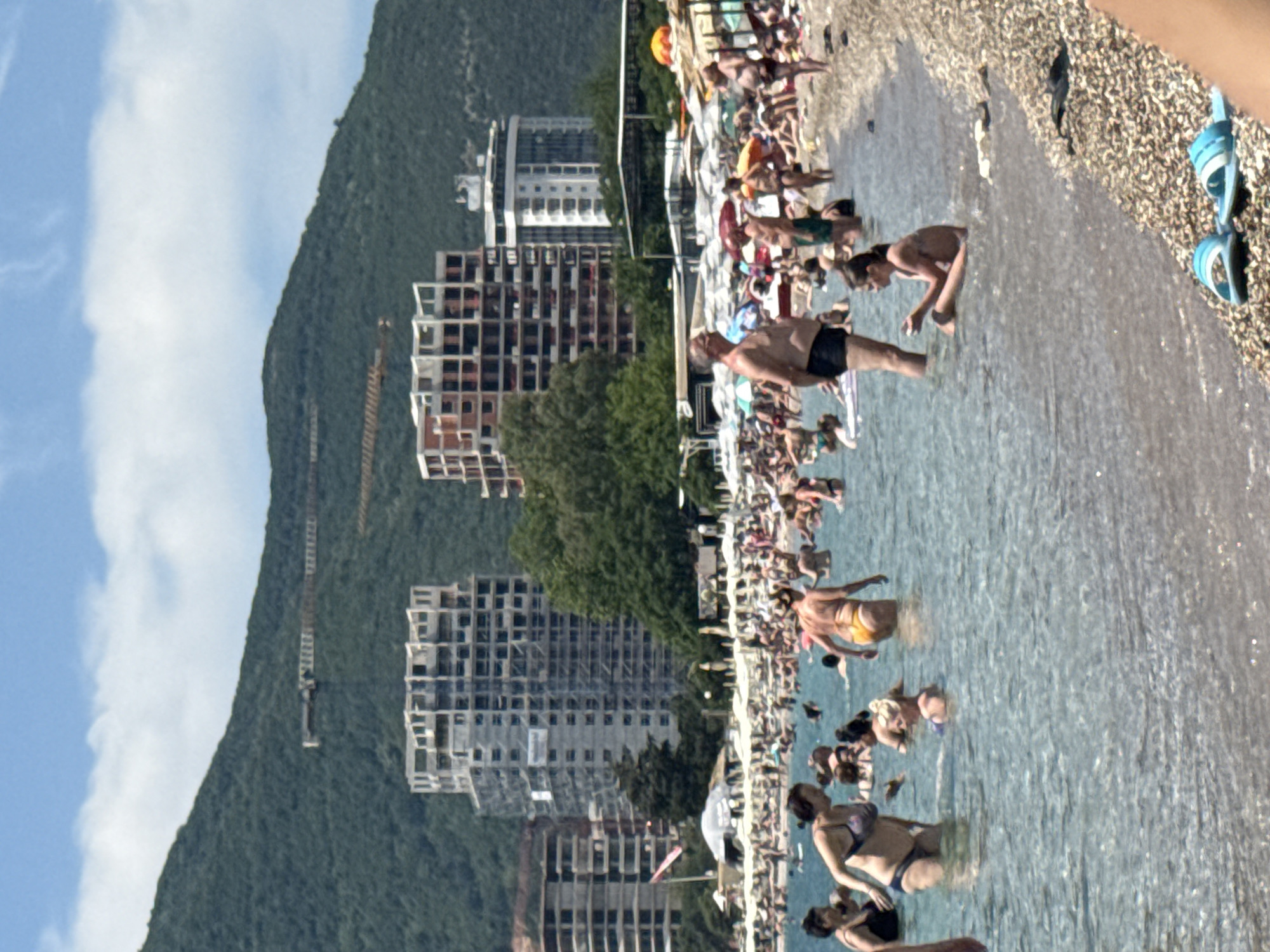 In the city of Boreti, Montenegro, a crowded beach scene unfolds under a bright, cloudy sky. The foreground features a pebbled shoreline with a pair of blue and white flip-flops. The shallow, clear water is populated by numerous individuals, primarily women and children, engaged in various activities. Some are wading, others are standing, and a few are further out. A woman in a dark swimsuit and a woman in a yellow bikini are prominent in the mid-ground, as is a young girl sitting in the water with her hands clasped. In the background, a bustling beach with many people sunbathing and relaxing under umbrellas stretches out. Beyond the beach, a row of modern, multi-story buildings, some appearing to be under construction, are visible. A lush, green, tree-covered mountain rises behind the buildings, with construction cranes visible on its slopes. The overall atmosphere is one of a lively summer day at a popular tourist destination.