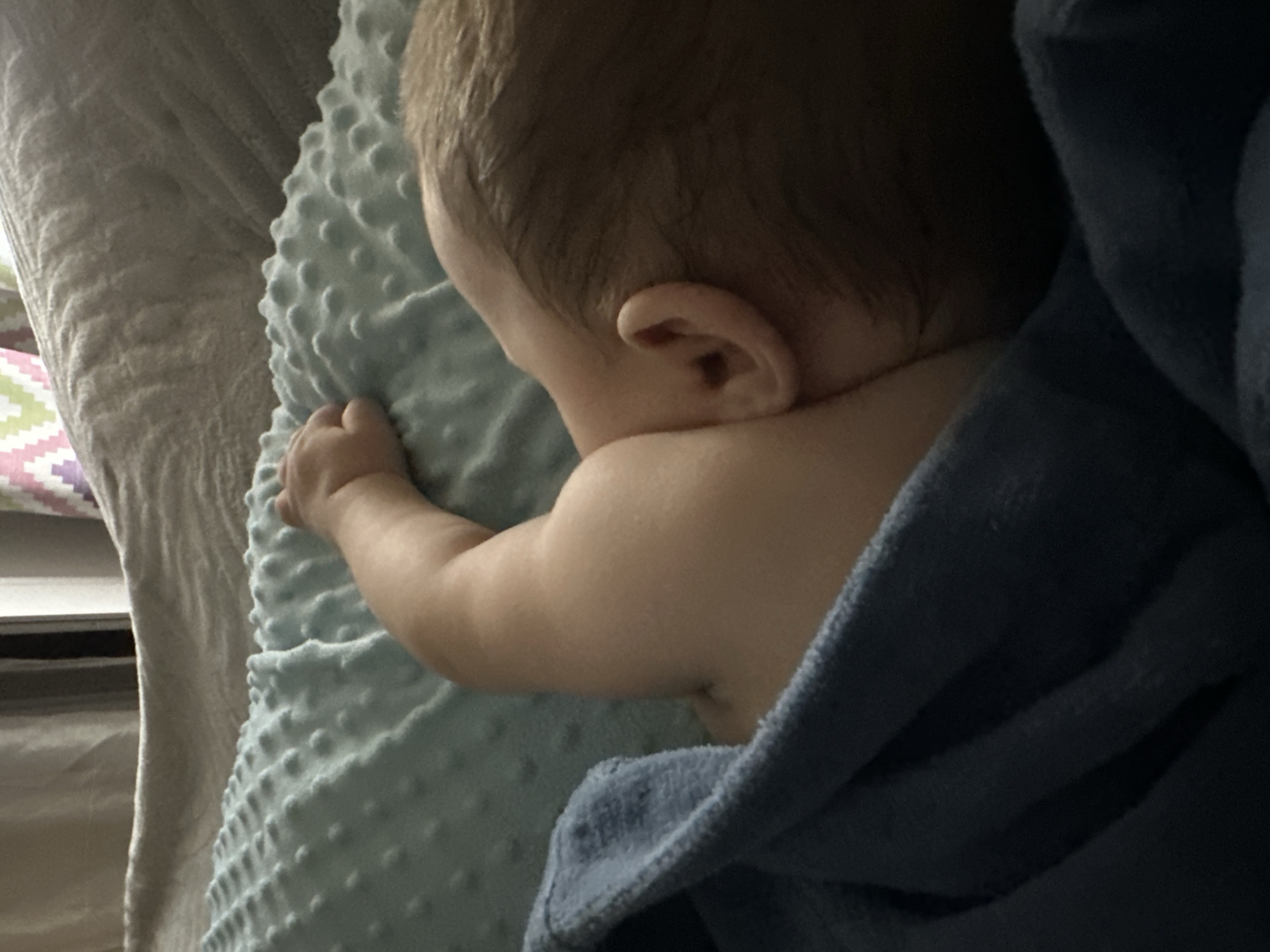 A baby lies on a light blue blanket with raised bumps, partially covered by a blue blanket. The baby's back, arm, and part of their head are visible, with a focus on their ear. The environment appears to be indoors, possibly in a home setting.