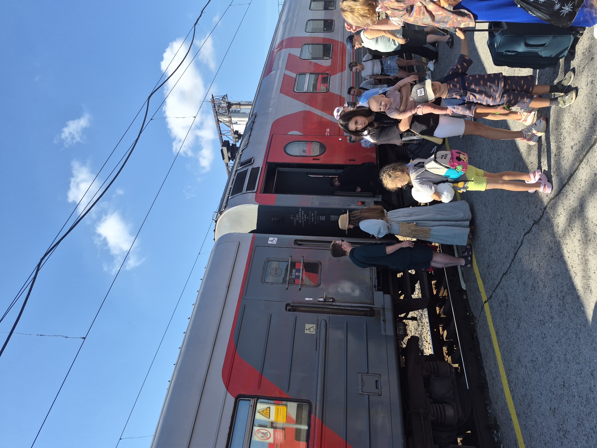 This image captures a scene at a train station platform in Yekaterinburg, Russia, on a sunny day. A train with a prominent red and grey livery, featuring the Russian Railways logo, is stopped at the platform with its doors open. Several people are visible, some boarding or disembarking the train, while others are waiting on the platform.
In the foreground, a young boy wearing a cap and a patterned shirt stands next to a dark blue suitcase, holding onto its handle. To his left, a young girl in yellow shorts and a grey t-shirt, carrying a backpack and a stuffed toy, stands facing away from the camera. Between them, a woman in a black t-shirt and white shorts holds a baby. Behind them, other individuals are visible, including a man in a dark shirt and shorts talking to a woman in a light blue dress and a straw hat. Further back, more passengers are scattered along the platform.
The platform itself is paved with asphalt, marked by a bright yellow line along the edge. The train tracks are visible below the platform. Overhead, electrical wires for the train are strung across the clear blue sky, with a few scattered white clouds. The overall atmosphere suggests a moment of travel or departure.