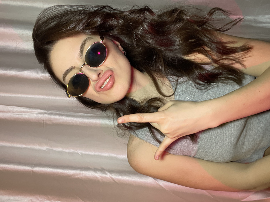A woman with long, wavy brunette hair poses on a pink satin sheet. She wears round sunglasses and a gray top. Her lips are pursed, and she holds up her hand in a gesture. The lighting is soft, with a pinkish hue.