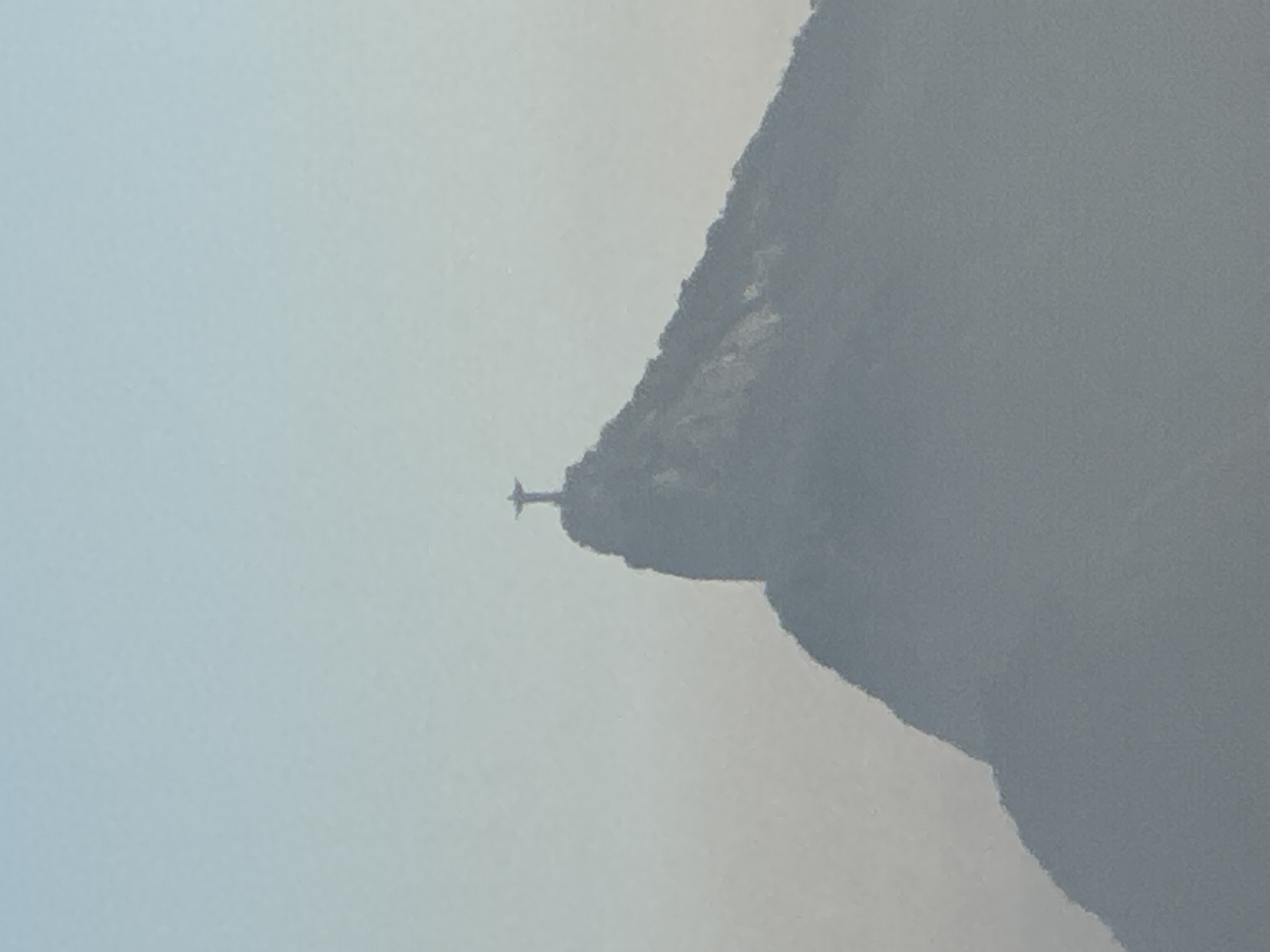 The image shows a distant, silhouetted view of the Christ the Redeemer statue atop Corcovado Mountain. The statue is positioned on the peak of the mountain, with its arms outstretched. The mountain itself is dark and appears to be covered in foliage, though the detail is obscured by haze. The background consists of a pale blue, hazy sky that gradually lightens towards the top of the frame. The scene is captured from a distance, with a noticeable atmospheric haze softening the details of both the statue and the mountain. This iconic landmark is located in Rio de Janeiro, Brazil, and is visible from various points in the city, including across Guanabara Bay in Niterói.