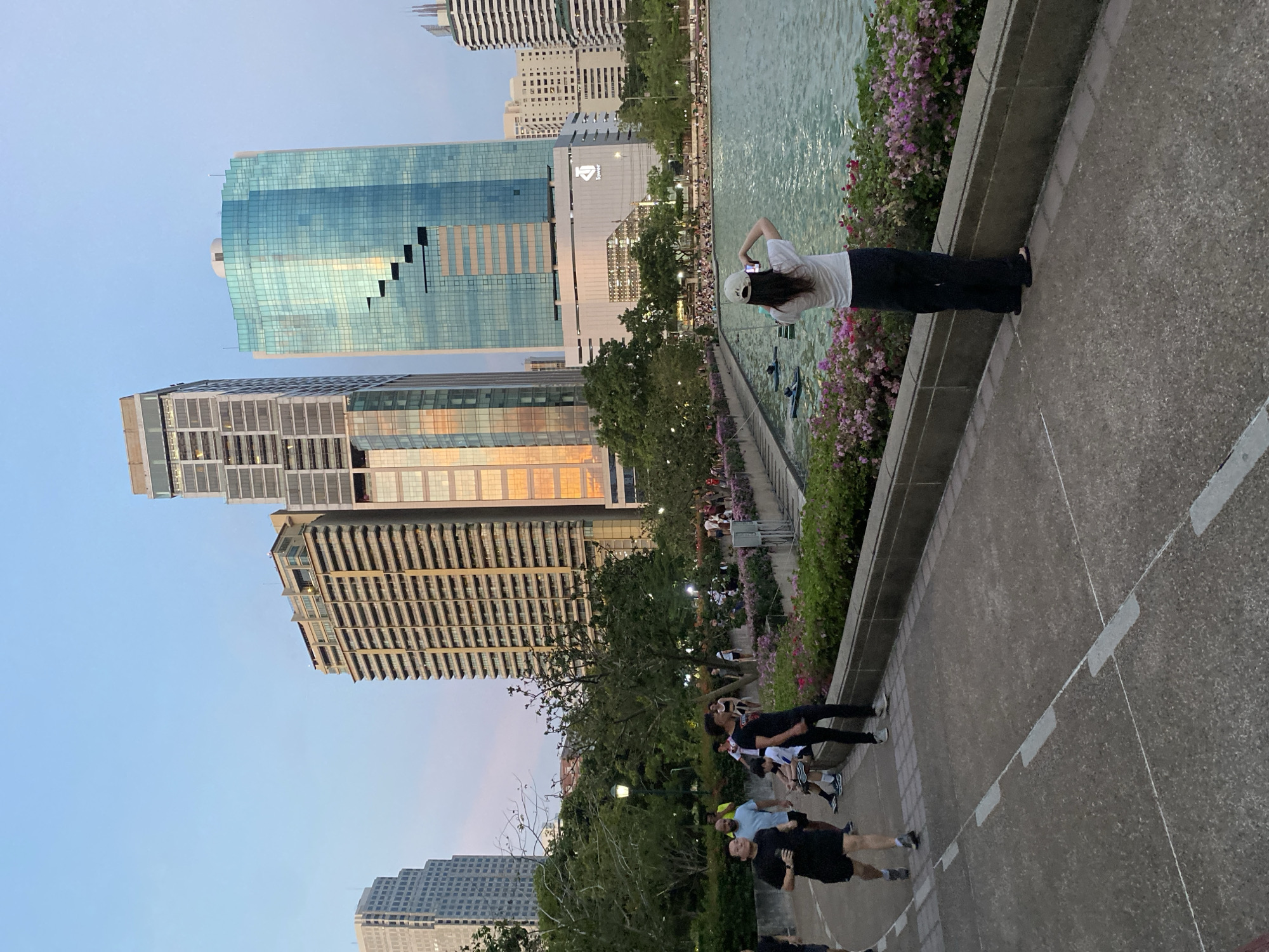 A street-level, medium-distance shot shows a partially obstructed view of the skyline of Bangkok, Thailand, on an overcast day. Multiple buildings dominate the horizon. In the foreground, a group of people is taking in the view. A woman in a white shirt and black pants stands at the edge of a paved area, taking a picture of the scene with her phone. A body of water with pink flowers is visible next to the woman. The paved area where the people are standing is marked with white lines. Trees and shrubs are visible in the midground, partially obscuring the buildings. The overall mood is relaxed and casual, suggesting a public space or park setting.