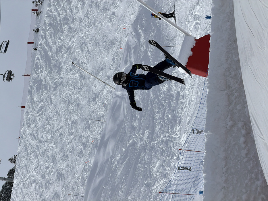 The scene depicts a snow-covered ski slope within the ski resort area of Les Allues, France. A single individual, dressed in a dark blue ski suit with light blue accents, black helmet, black gloves, and white-framed goggles, is captured mid-air on skis. The skier is positioned left of center, airborne over a red, snow-covered pyramidal base for a white pole, which is anchored in the snow. Their body is crouched with arms extended, and their skis, marked "K2," are parallel to the ground. A blue safety net fence is visible along the lower left quadrant of the image, running horizontally above a snow bank. In the background, a heavily tracked ski slope recedes uphill. Two empty chairlift chairs are visible against a light sky in the upper right. Multiple red marker poles dot the slope. To the far right, partially obscured by the main subject, another skier in a yellow jacket and dark pants is visible further down the slope. Sparse evergreen trees are visible in the far upper left background.