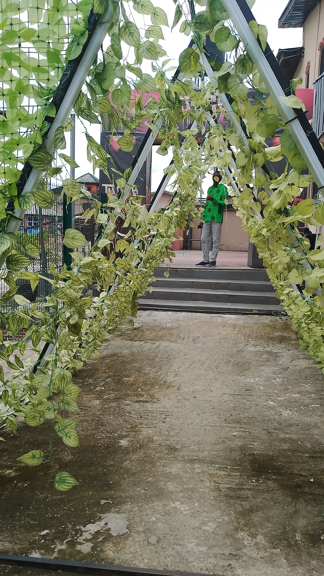 The photo depicts a person standing at the top of a set of stairs under an archway of greenery. The person is wearing a green patterned shirt and grey pants. The archway is composed of a metal structure covered in artificial green vines. The flooring in the foreground appears to be concrete with visible wear and discoloration. In the background, parts of buildings can be seen, including a fence, sections of painted walls, and upper floors with balconies or railings. The location is in the city of Unknown, Unknown.
